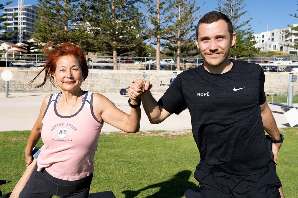 An older woman and a younger man outdoors in a sunny park, engaging in an arm wrestling match, smiling, with trees and buildings in the background.