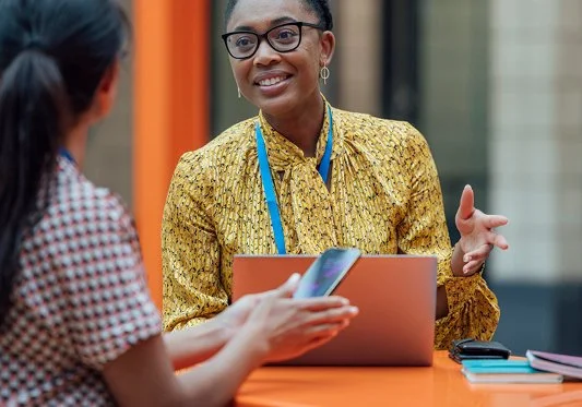 Two women engaging in a conversation at a meeting or conference, with one woman smiling and gesturing, and the other woman holding a phone.