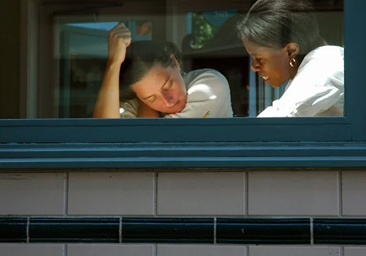 Two women looking through a window, one resting her head on her hand and the other engaging in conversation.