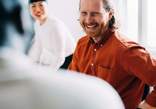 A man with long hair and a beard, wearing an orange shirt, smiling in a bright indoor setting with two women in the background.