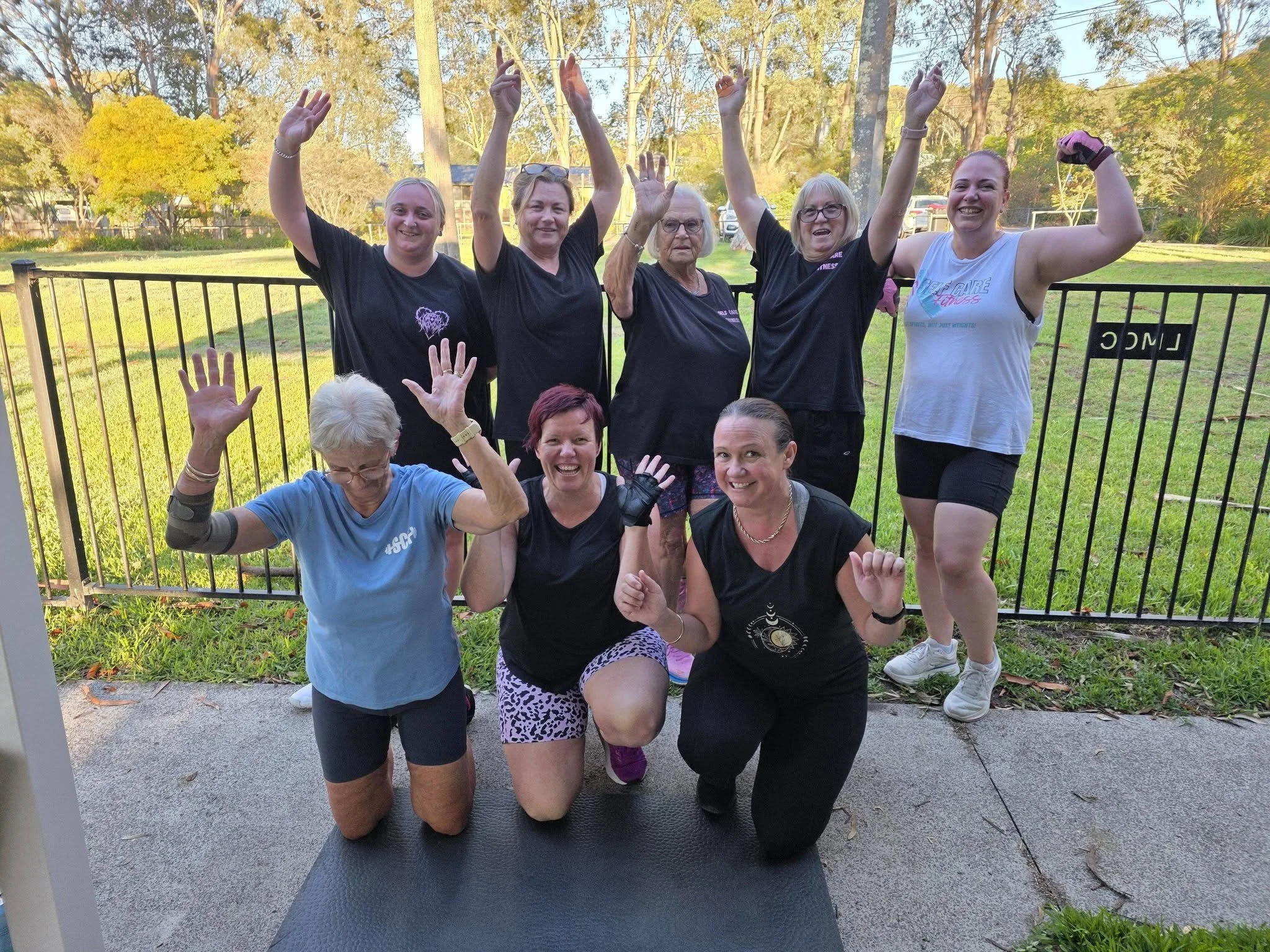 Group of six women and one person with gray hair and glasses, posing outdoors for a photo during a fitness or exercise session. They are making flexing or victory gestures with their arms. The women are smiling, and some are kneeling while others are standing behind a black metal fence. The background shows trees and a grassy park area with sunlight filtering through.