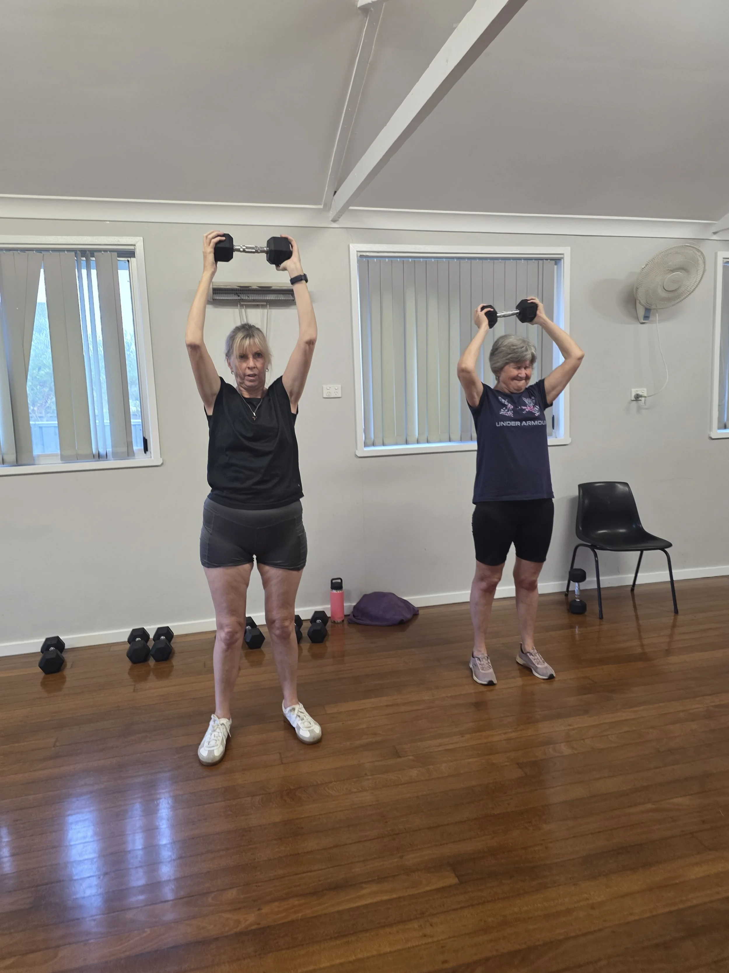 Two women are doing an exercise class by lifting dumbbells overhead in a room with wooden floors, white walls, and window blinds. The woman on the left is wearing a black shirt and shorts, and the woman on the right is wearing a navy shirt and shorts. There are multiple dumbbells on the floor and a chair with a water bottle nearby.