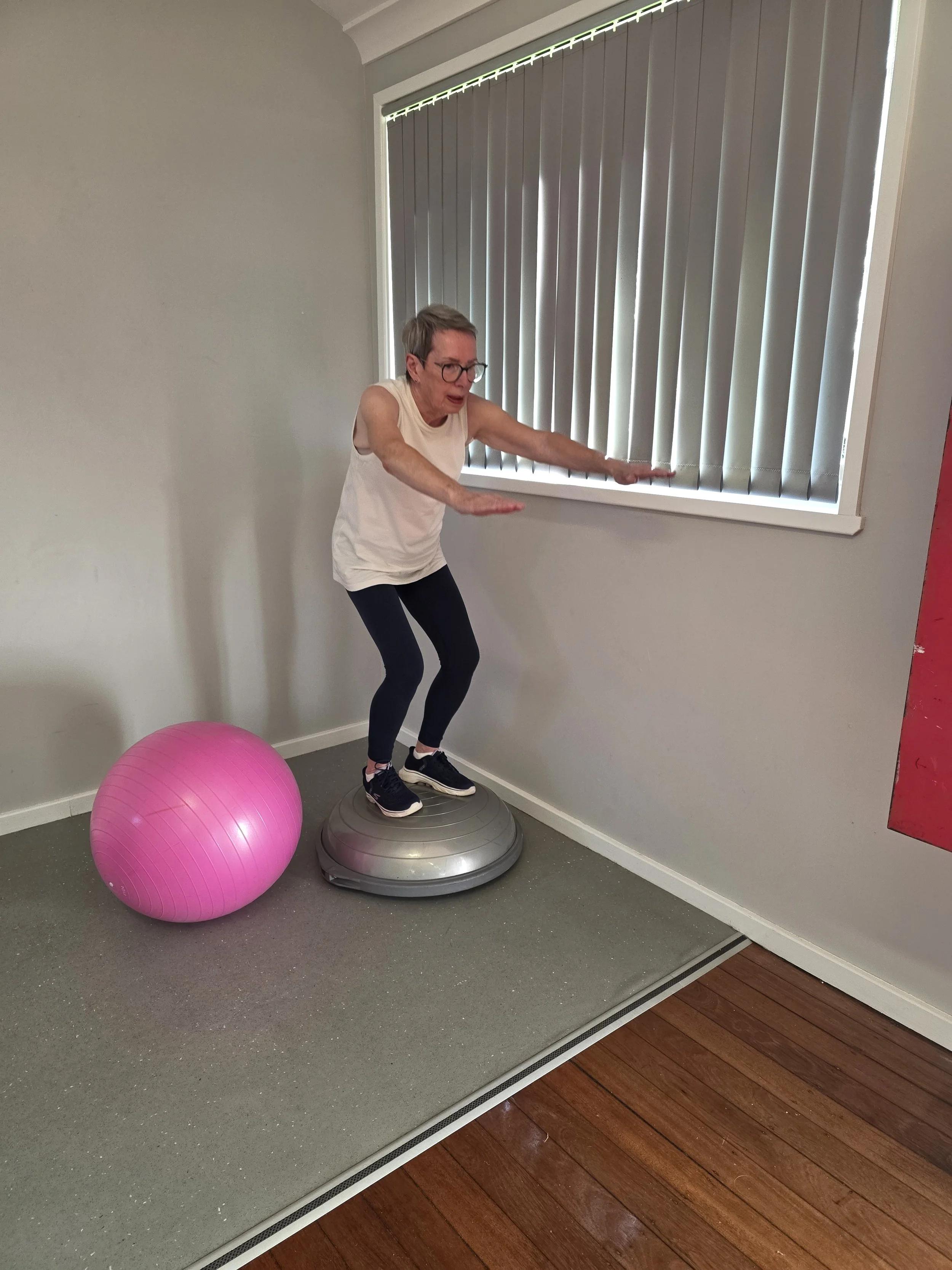 A woman with short gray hair and glasses balancing on a gray exercise balance disc while extending her arms forward, with a large pink exercise ball nearby, in a room with gray walls, a window with vertical blinds, and a wooden floor.