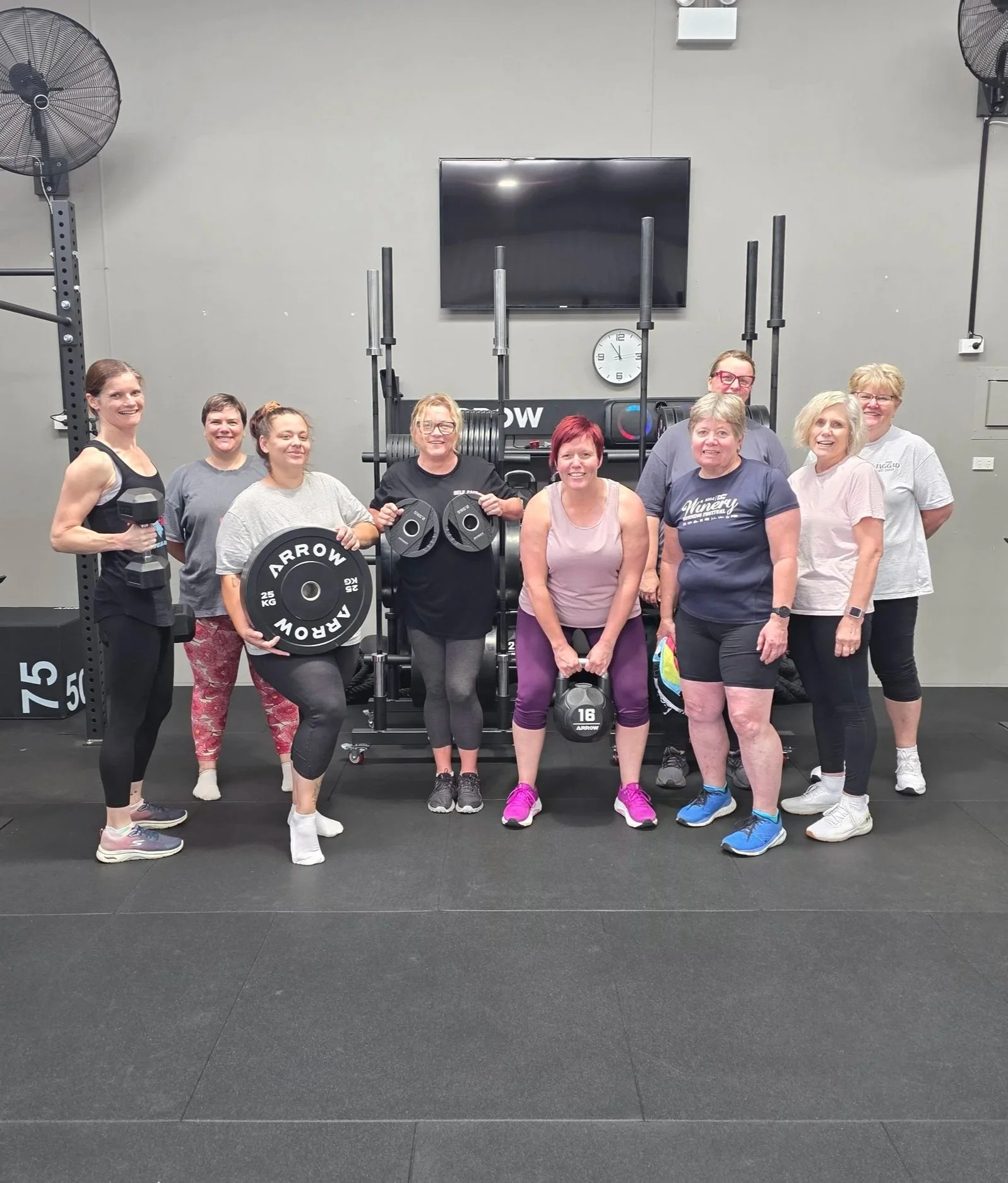 Group of nine women posing in a gym, some holding weights and standing in front of gym equipment. The women are smiling and dressed in workout clothes.