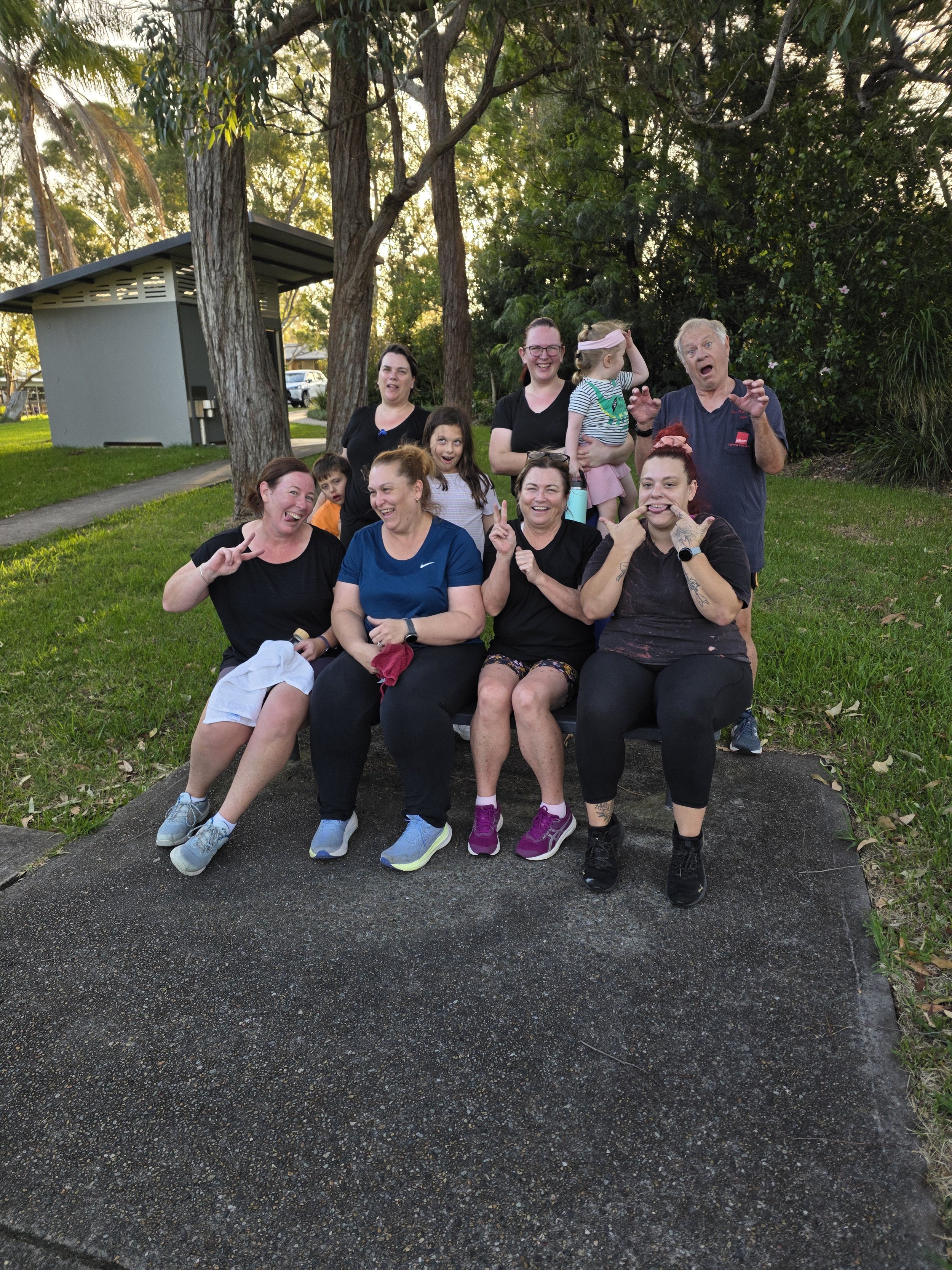 A group of ten people, including adults and children, are gathered outdoors in a park-like setting, sitting and standing on a bench and grass, with trees and a small building in the background, enjoying a sunny day.