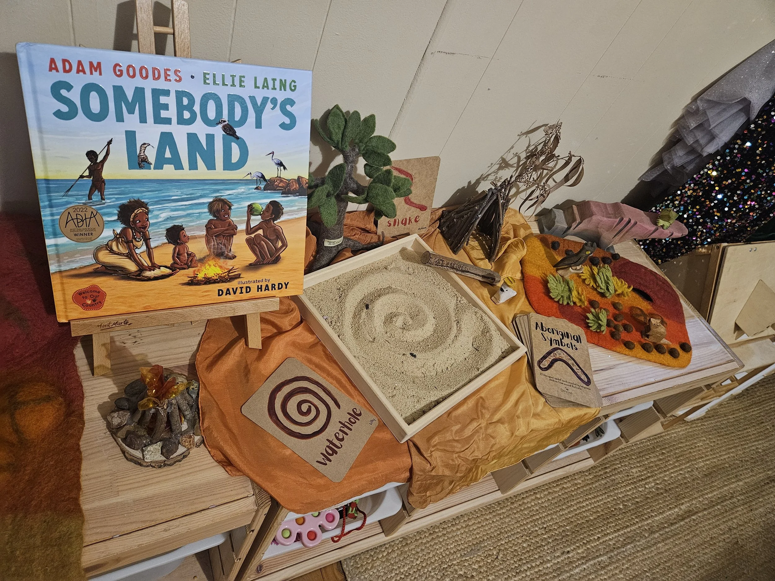 Child exploring an Indigenous-themed picture book alongside loose parts and sensory materials during a NAIDOC Week inspired play activity.