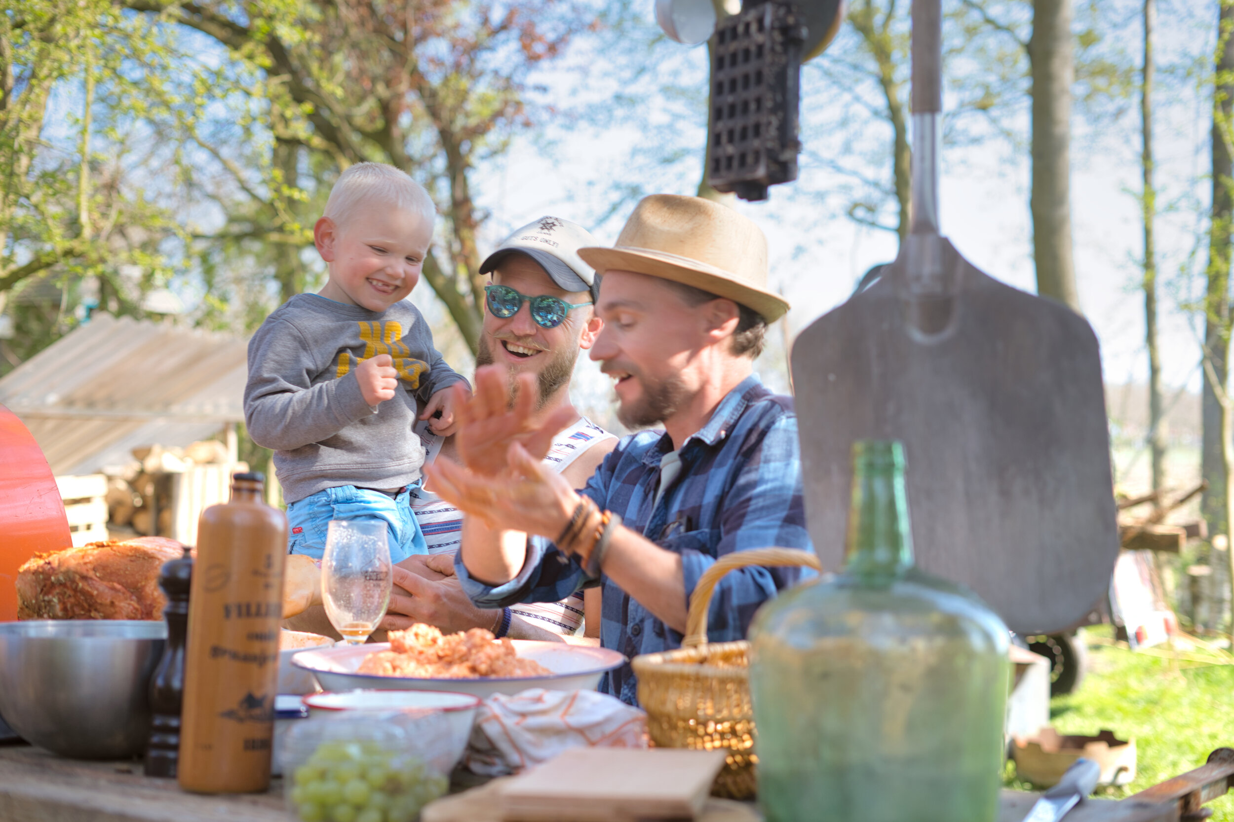 Twee mannen met een kind genieten van een picknick buiten aan een tafel met voedsel en drank, op een zonnige dag.