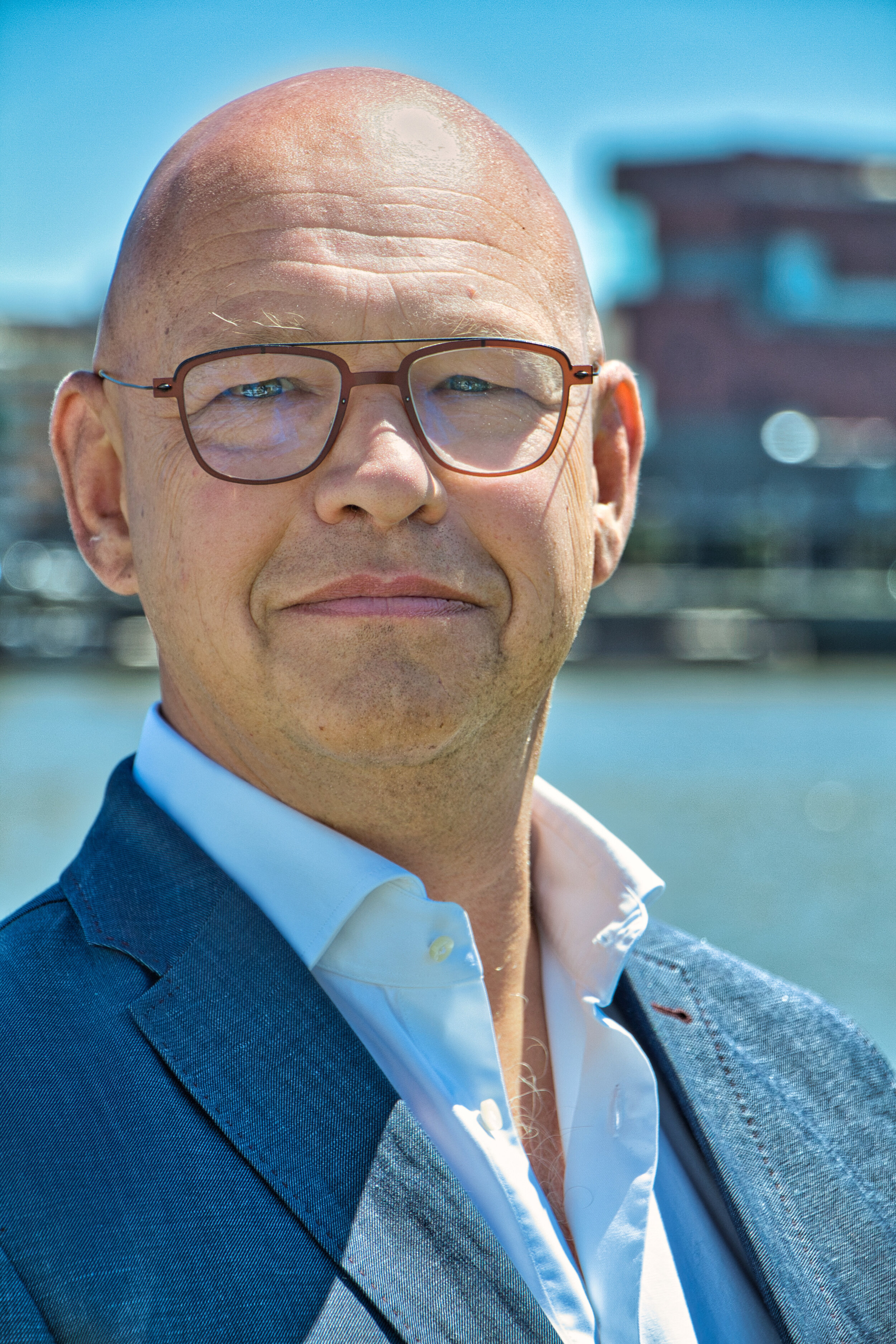 Portret van een kale man met bril en een blauw pak, buiten in de zon met wazige stadsachtergrond.