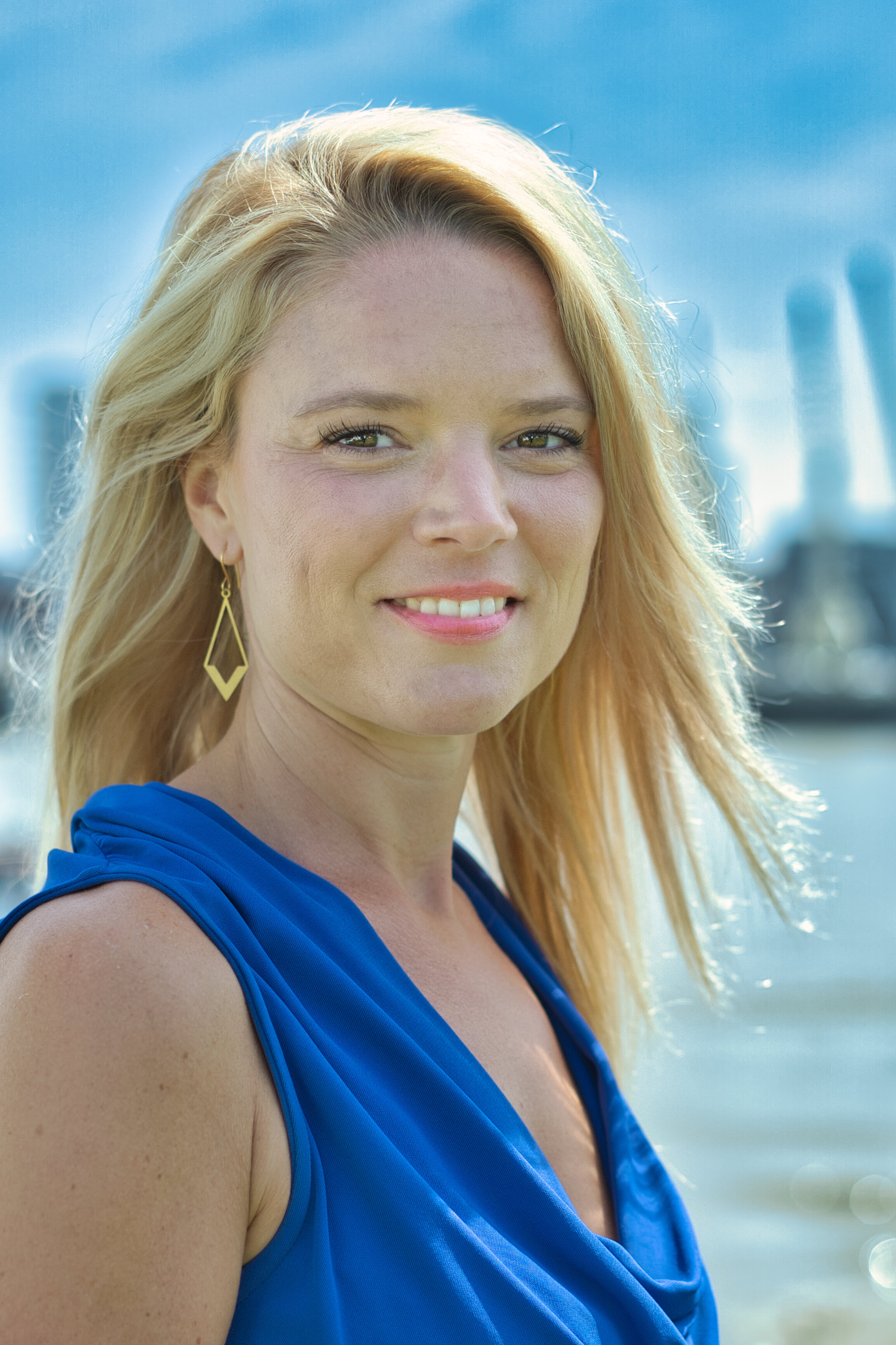 Blonde vrouw met blauwe jurk en goudkleurige oorbellen buiten bij water.