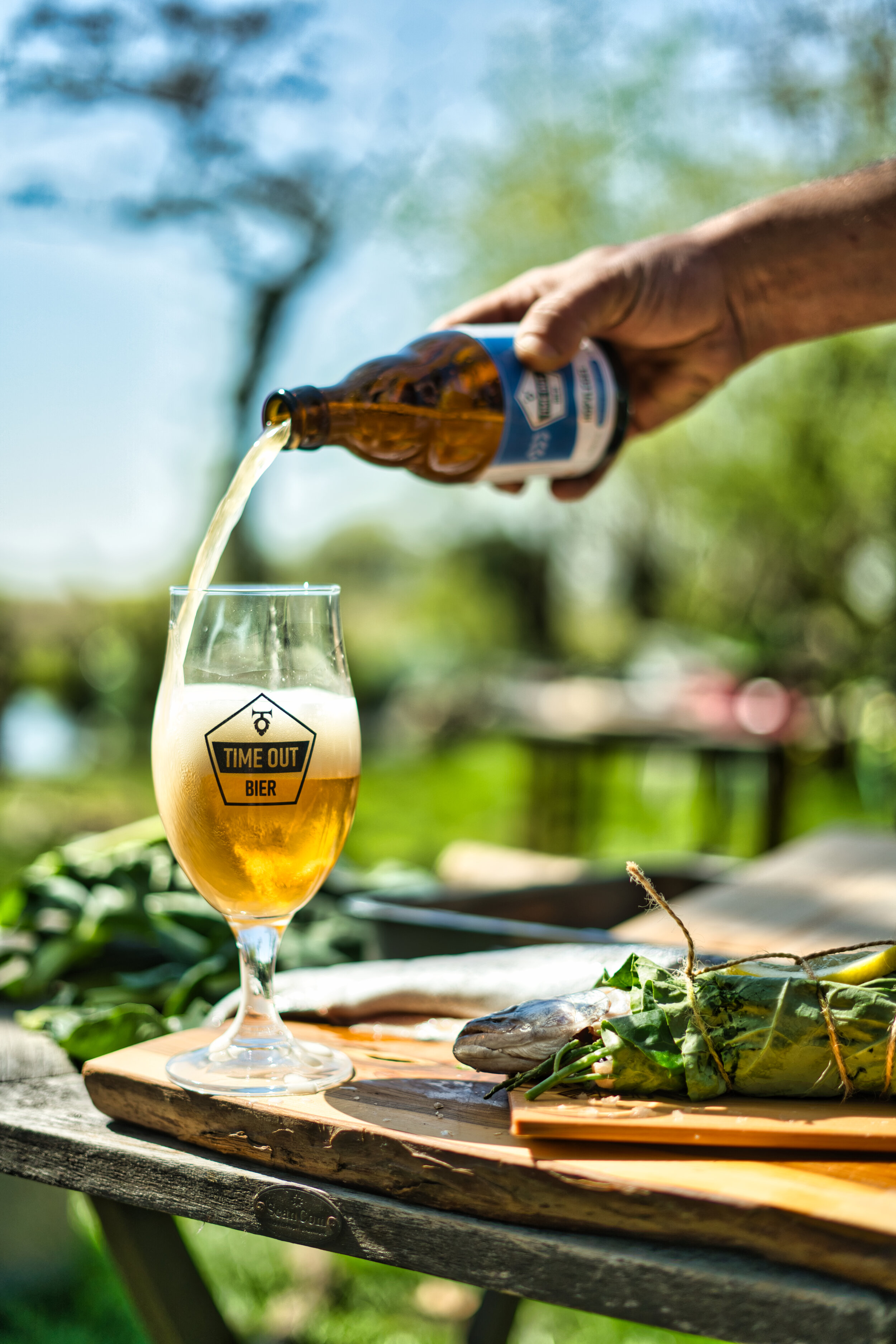 Een hand schenkt bier in een glas met het opschrift 'TIME OUT BIER' op een zonnige dag buiten. Op de voorgrond ligt een vis gewikkeld in bladeren op een houten plank.