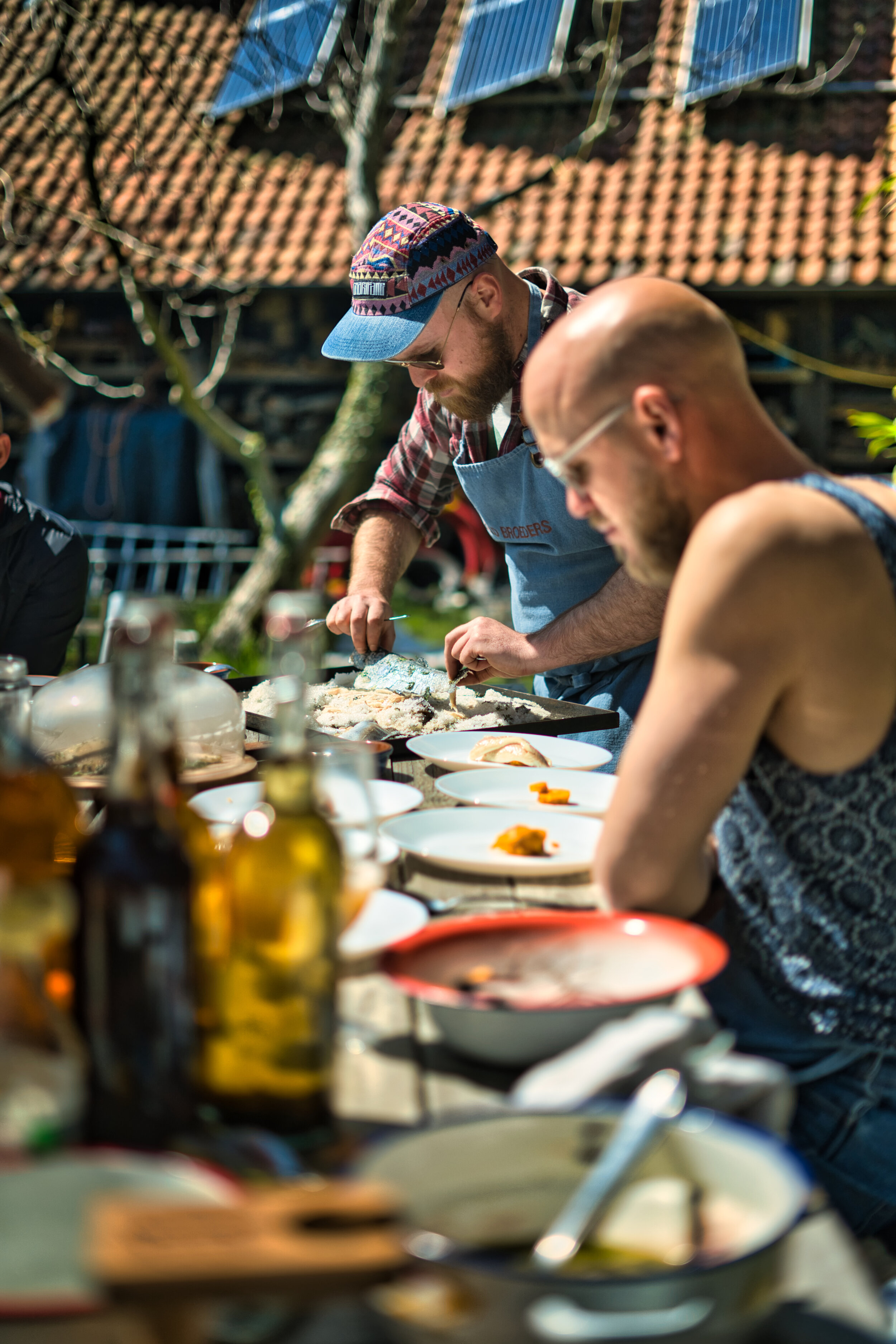 Mensen bereiden eten buitenshuis bij een tafel vol gerechten en ingrediënten, onder een zonnige hemel met zonnepanelen op de achtergrond.