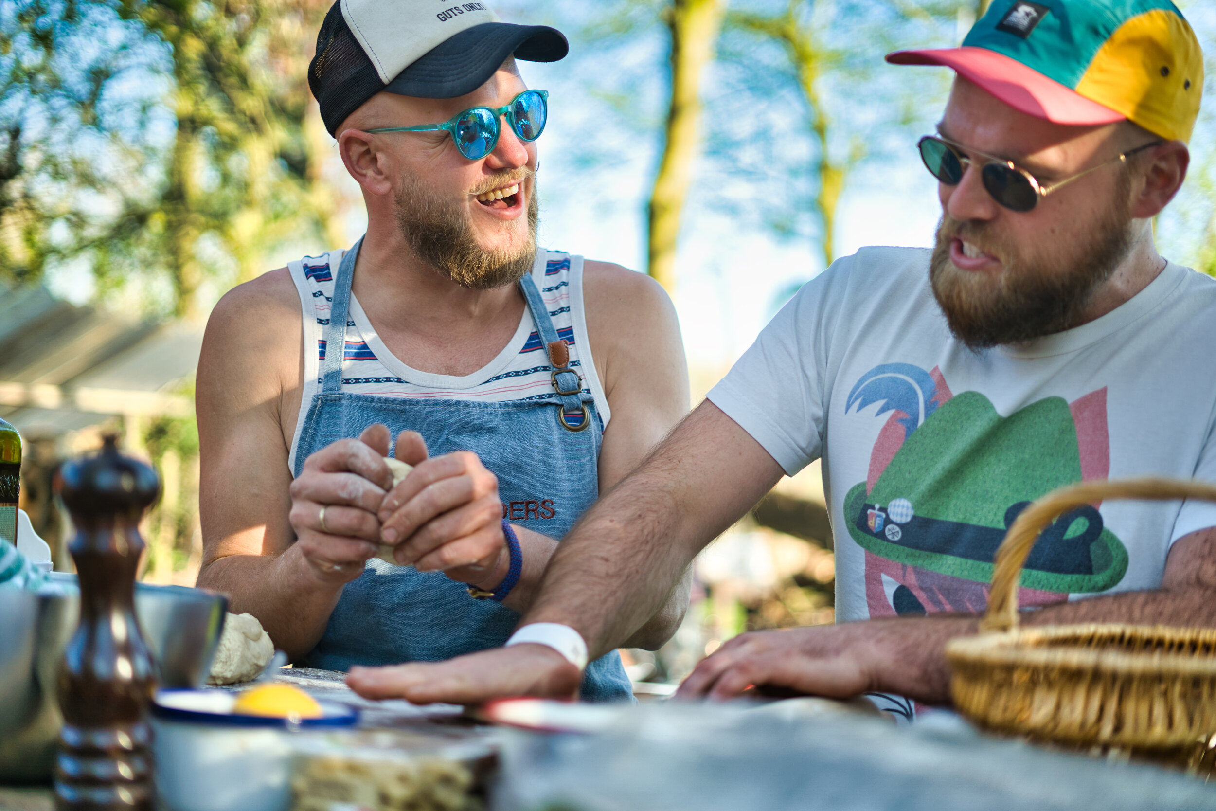 Twee mannen zitten buiten aan een tafel, bezig met voedselbereiding. Ze dragen petten en zonnebrillen. Er zijn verschillende kookbenodigdheden en een mand op tafel. De sfeer is ontspannen en vrolijk.