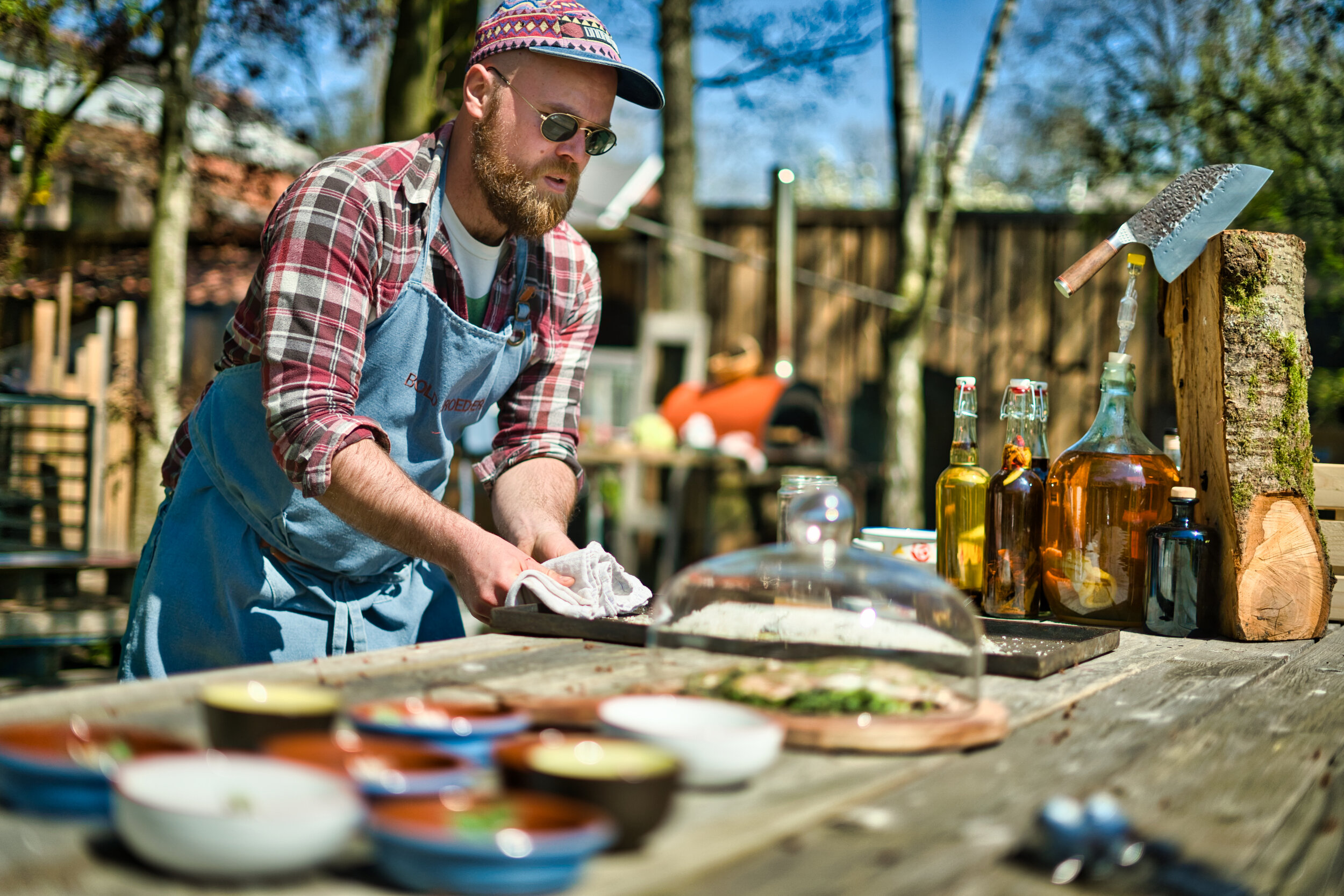 Man in schort aan het koken buiten, met diverse kommen en flessen op een houten tafel.