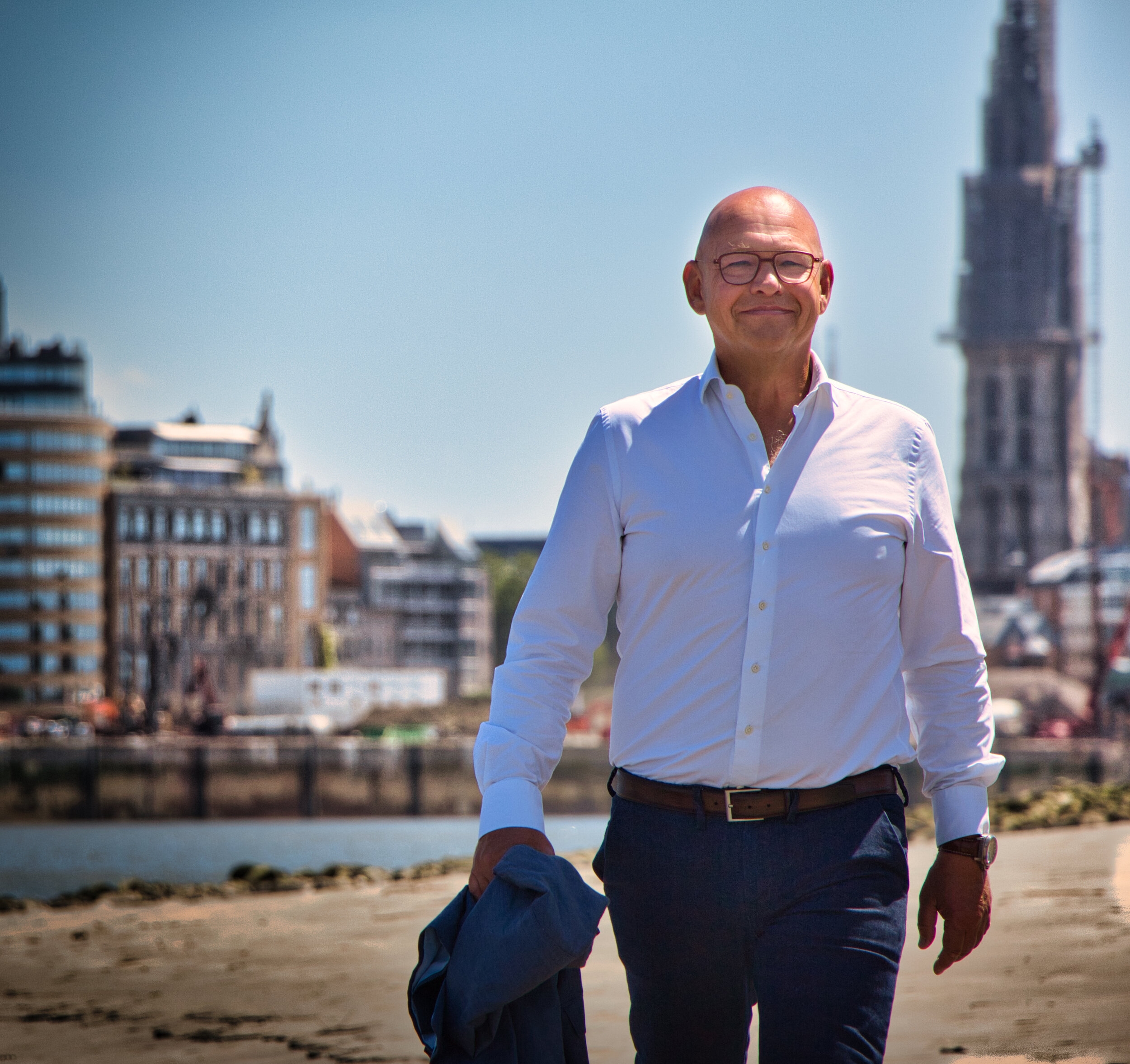 Man in een wit overhemd met een jasje in zijn hand loopt langs een waterkant, met stedelijke architectuur en een toren op de achtergrond.