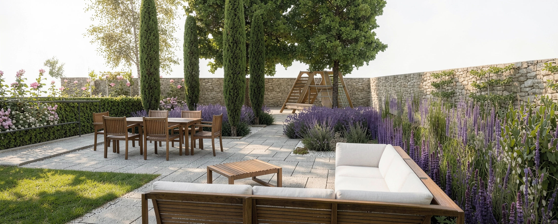 A landscaped backyard with a stone patio, wooden table and chairs, white sofa, tall cypress and deciduous trees, and purple flowering plants, enclosed by a stone wall.