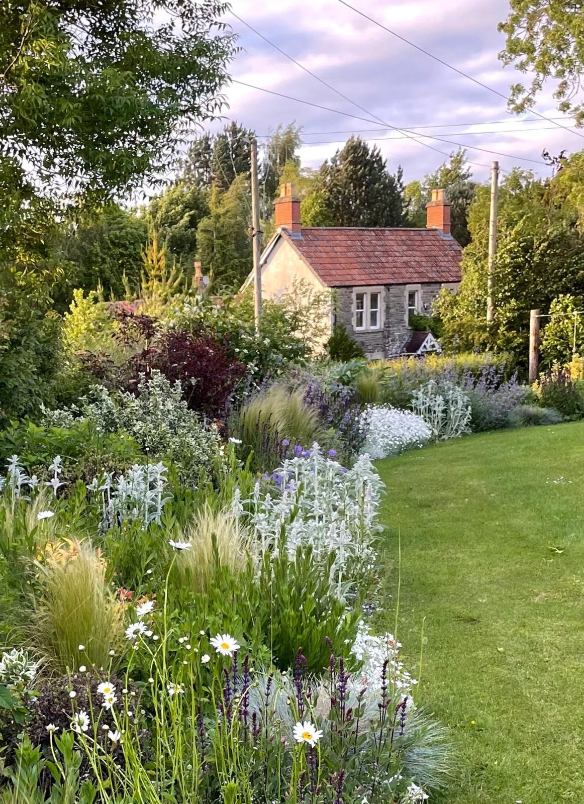 A garden with colorful flowering plants, a lush green lawn, and a small stone house in the background with red chimneys and a tiled roof, under a partly cloudy sky.