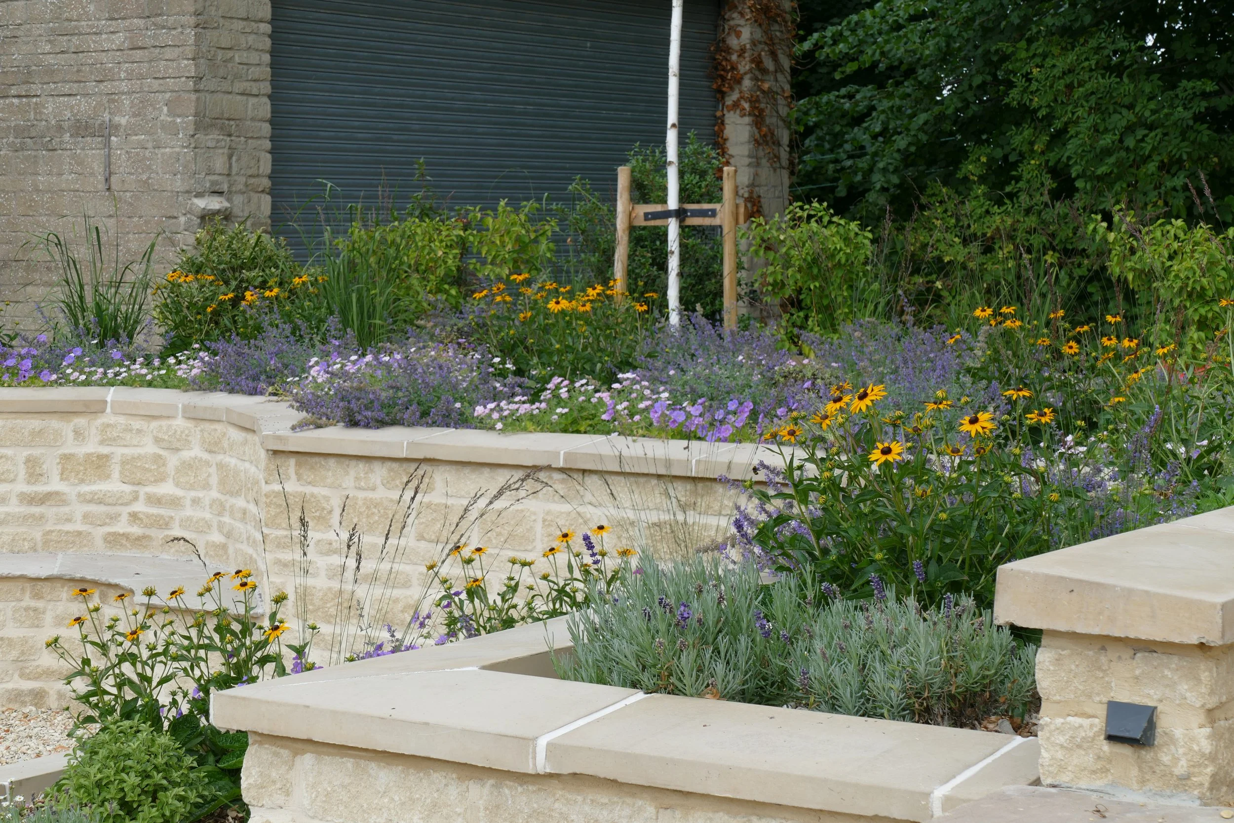 A garden with blooming flowers, concrete stairs, and a brick wall with a closed metal shutter door, surrounded by green foliage.