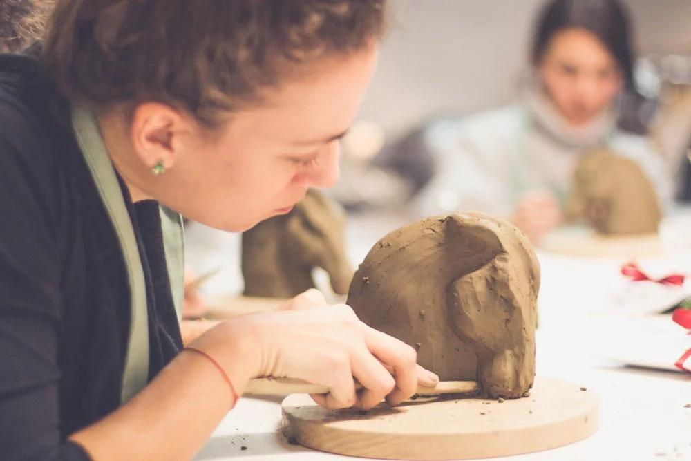 A woman sculpting a small animal figure out of clay, with another person in the background also working on clay art.