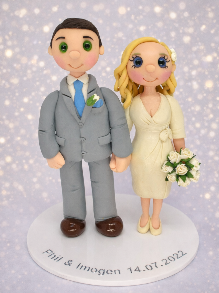 Bespoke wedding cake topper with bride and groom standing holding hands, groom in grey suit and bride in cream coloured dress holding a bouquet of flowers. 