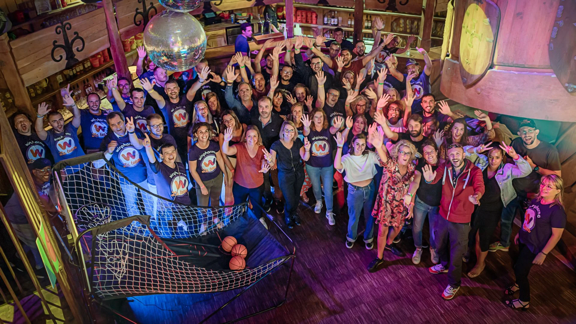 Un groupe de personnes souriantes et saluant, réunies dans un espace intérieur en bois, avec un ciel étoilé peint ou une lumière colorée. La majorité portent des t-shirts noirs avec un logo coloré, et il y a une ambiance festive et conviviale.