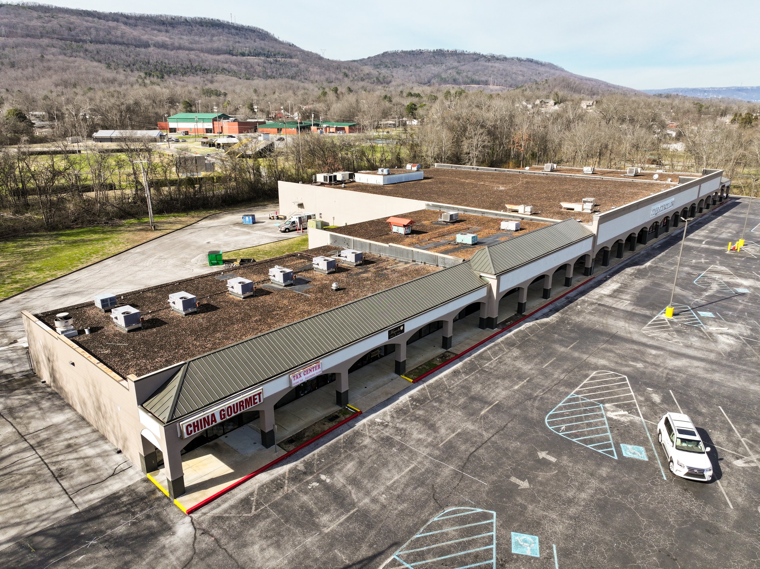 A shopping center with a sign that reads China Gourmet, in a mostly empty parking lot with a white car. The building has a brown roof and at least two sections, with a retail or restaurant storefront. Surrounding the shopping center are some empty parking spaces, green and blue dumpsters, and in the background, a wooded hillside and distant buildings.