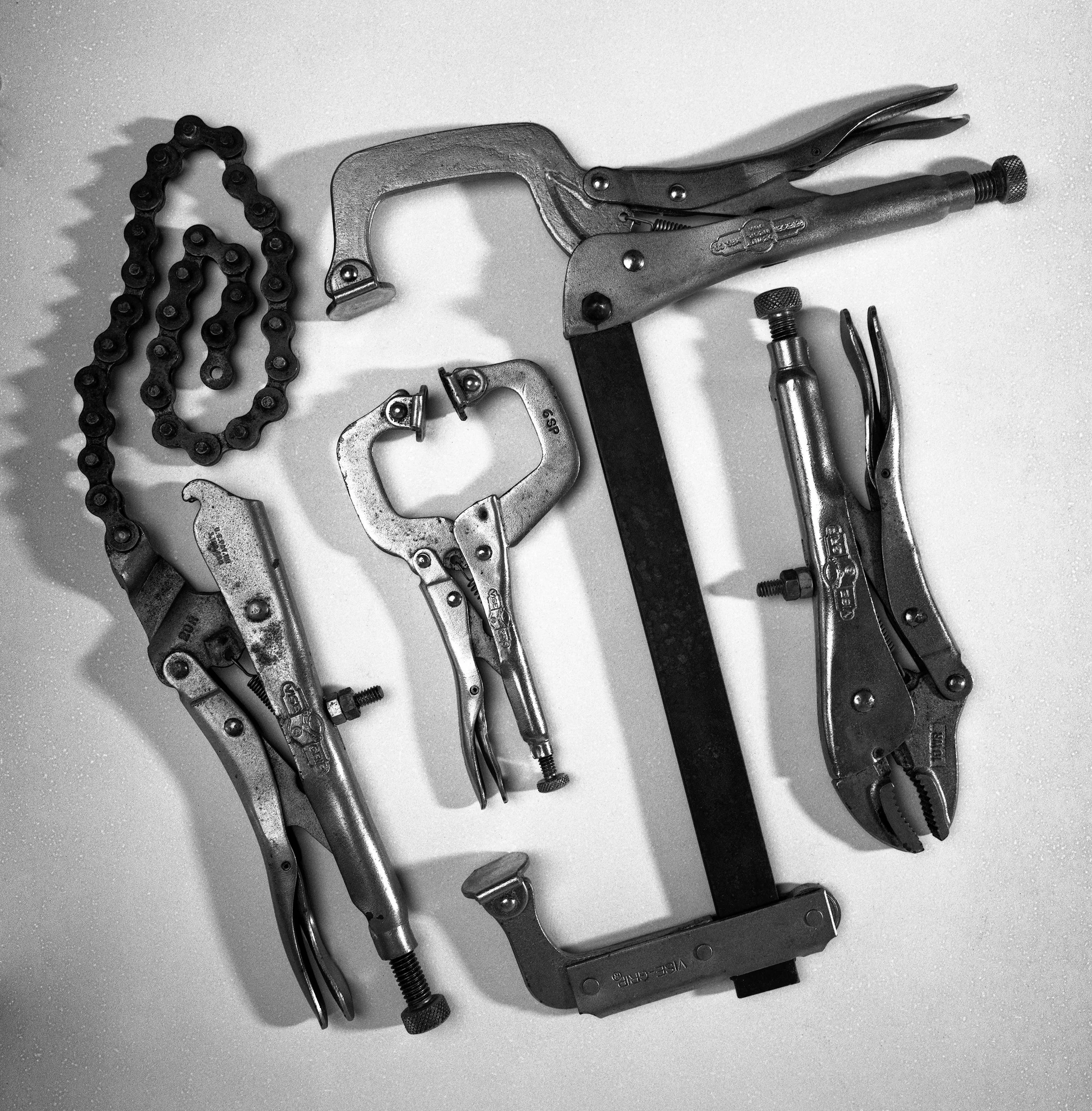 Collection of metal clamps, pliers, a chain, a C-clamp, and a saw on a white surface.
