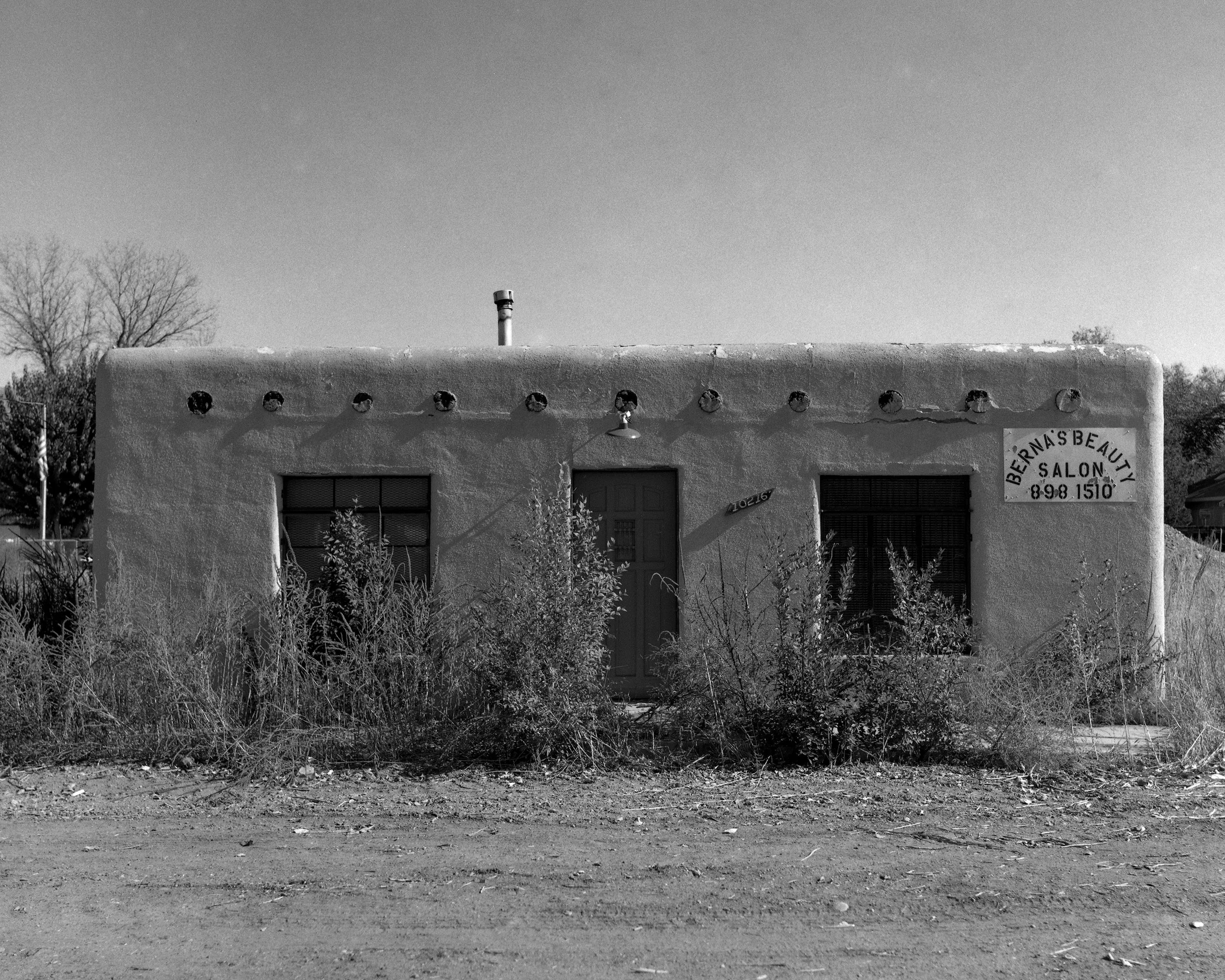 An old, abandoned building with a sign for Bernas Beauty Salon and overgrown weeds in front.