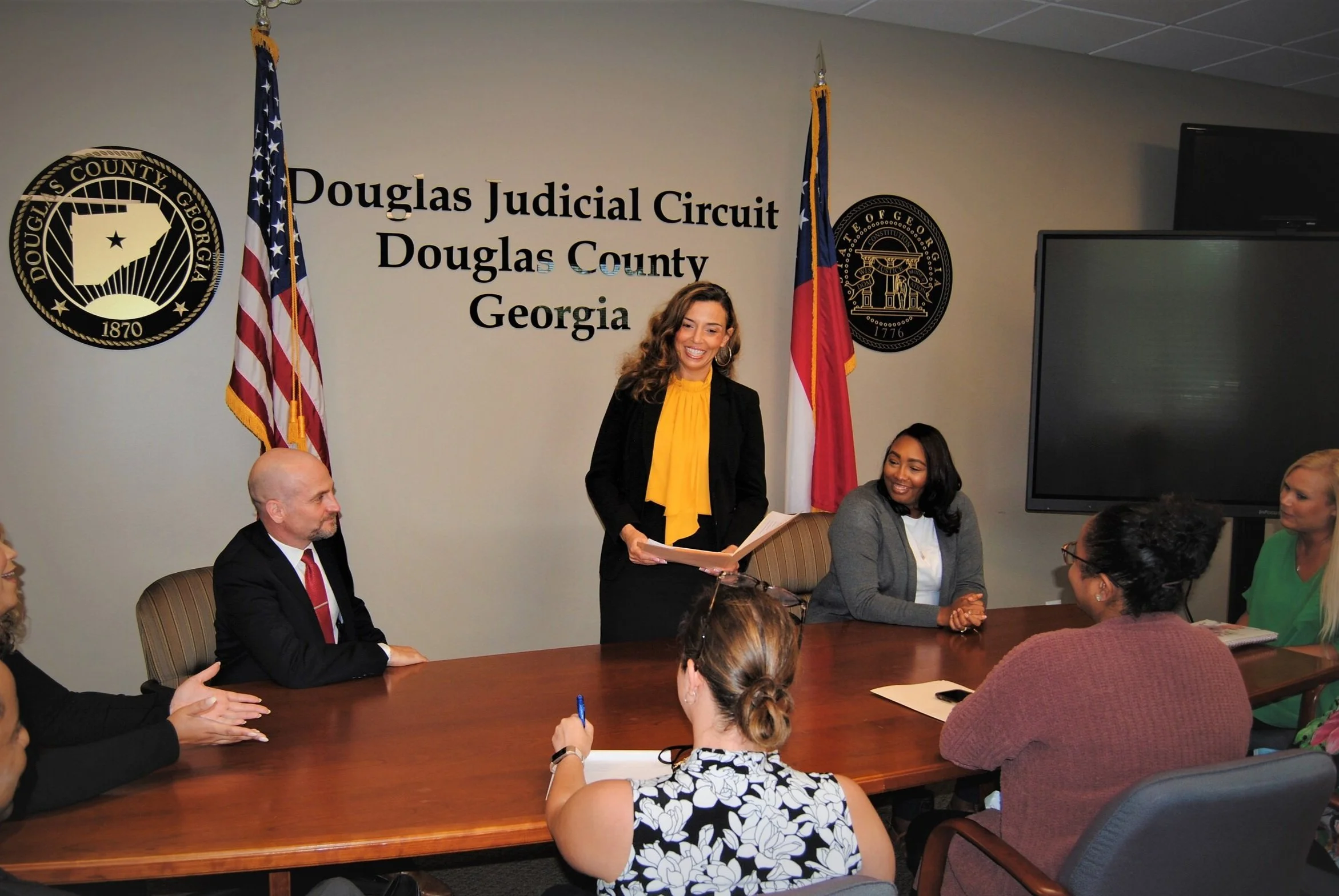 A woman in a black suit with a yellow top is standing at a table of five individuals. She is hold a file folder in her hand that is open.