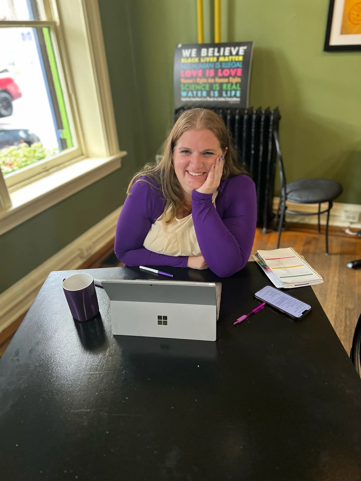 Katie wearing a cream blouse and purple sweater, leaning on a table with a computer in front of her and smiling.