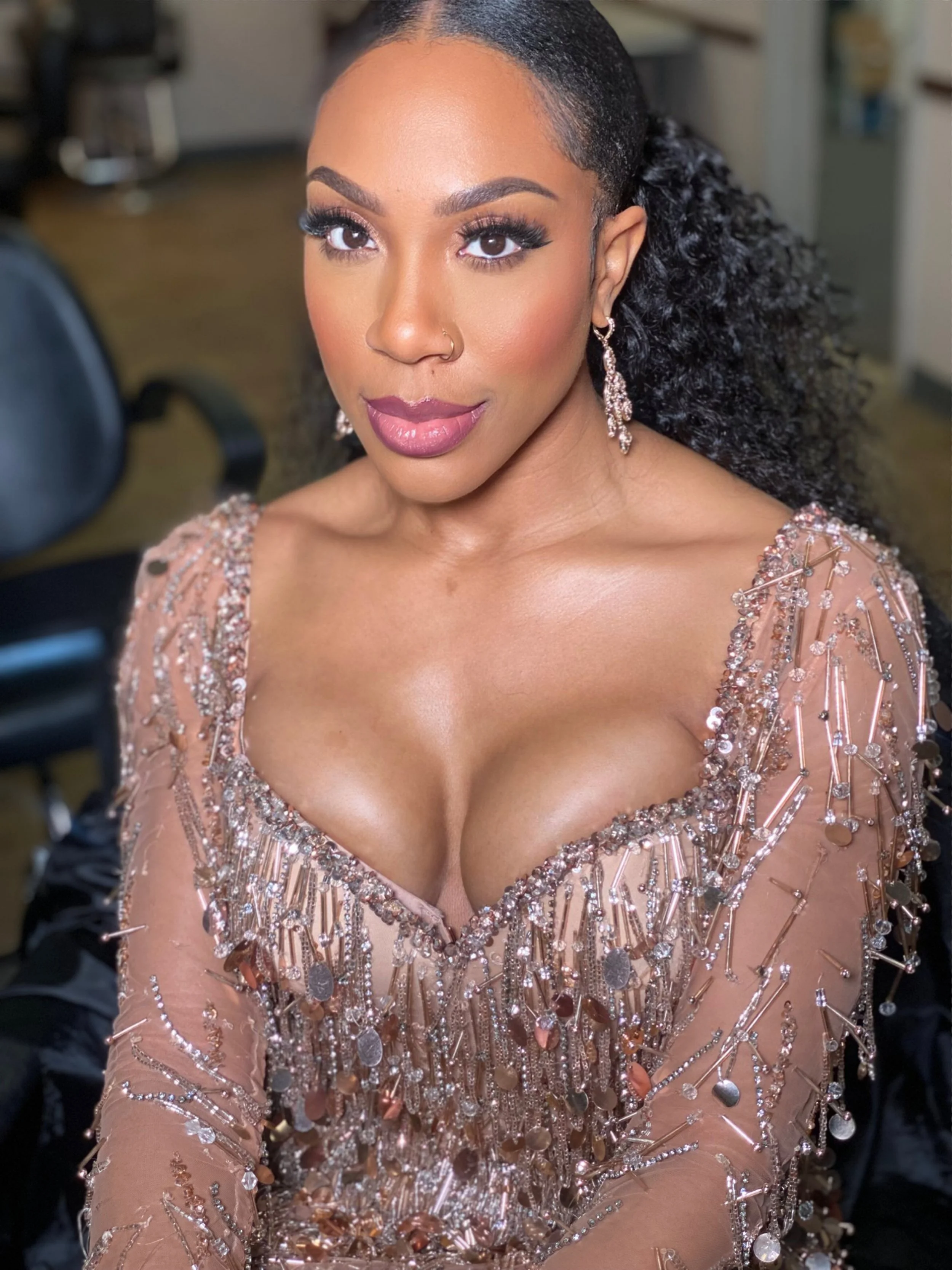 A woman with a confident expression dressed in an embellished, nude-colored gown with sequins and jewelry, posing in an indoor setting.