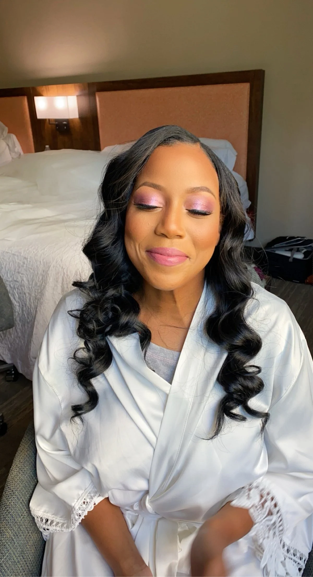 A woman with long, curly black hair and closed eyes, wearing pink eyeshadow and pink lipstick, sitting in a hotel room with a bed and headboard in the background.