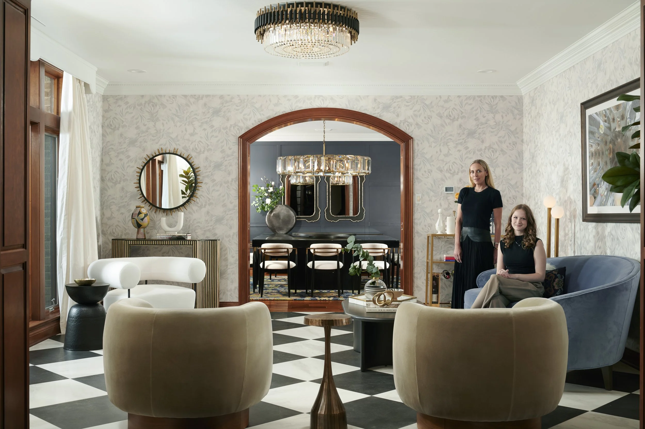 Living room with black and white checkered floor, cream and blue furniture, two women, one sitting on a blue sofa, one standing, elegant decor, large mirror, chandelier, and artwork.