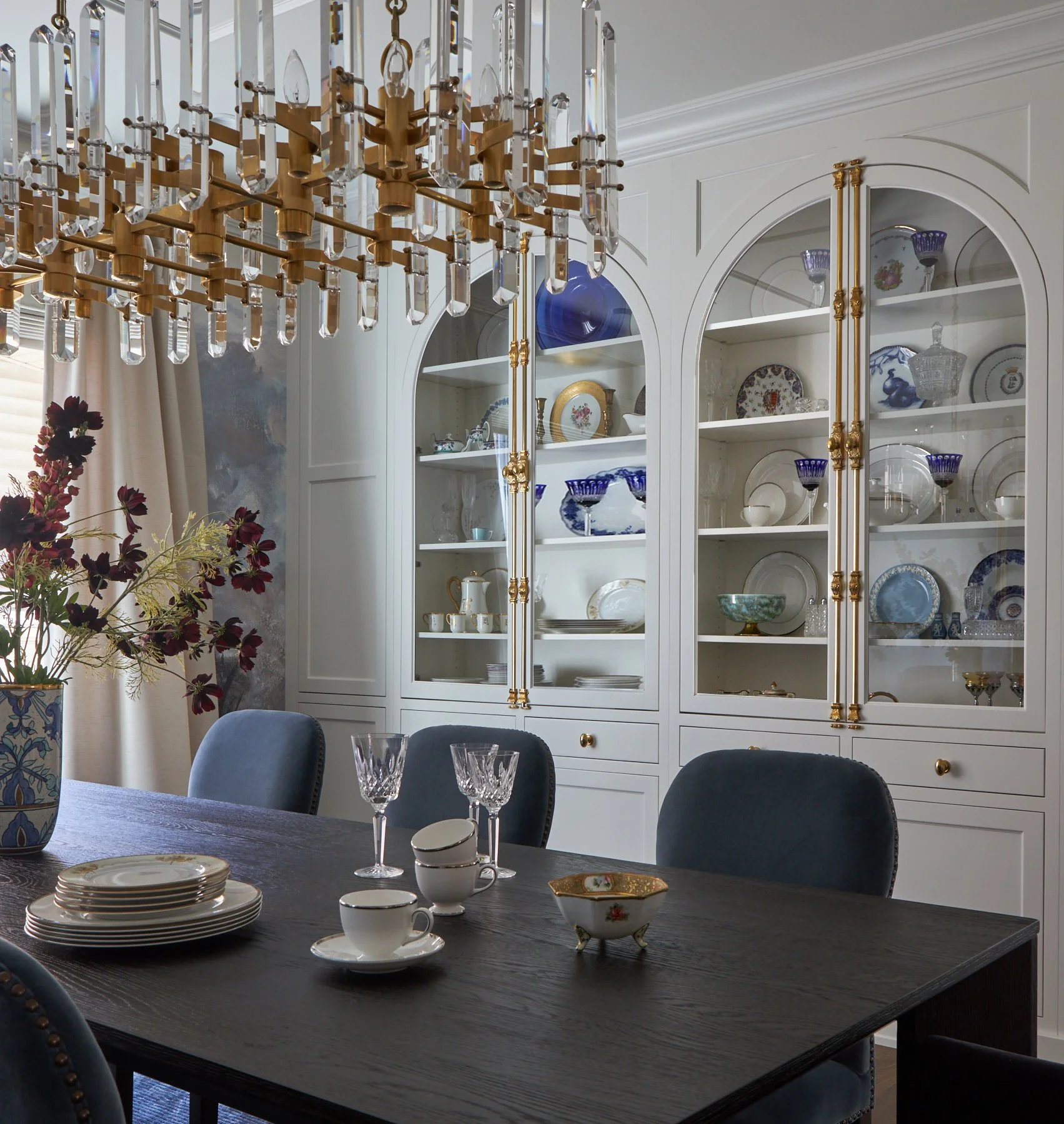 A dining room with a dark wooden table set with dishes, cups, and glasses. There are four upholstered chairs around the table. A white cabinet with gold accents and glass doors displays china, glassware, and decorative items. A large chandelier with crystal and gold details hangs above the table. A floral arrangement with dark red flowers is on a side table.