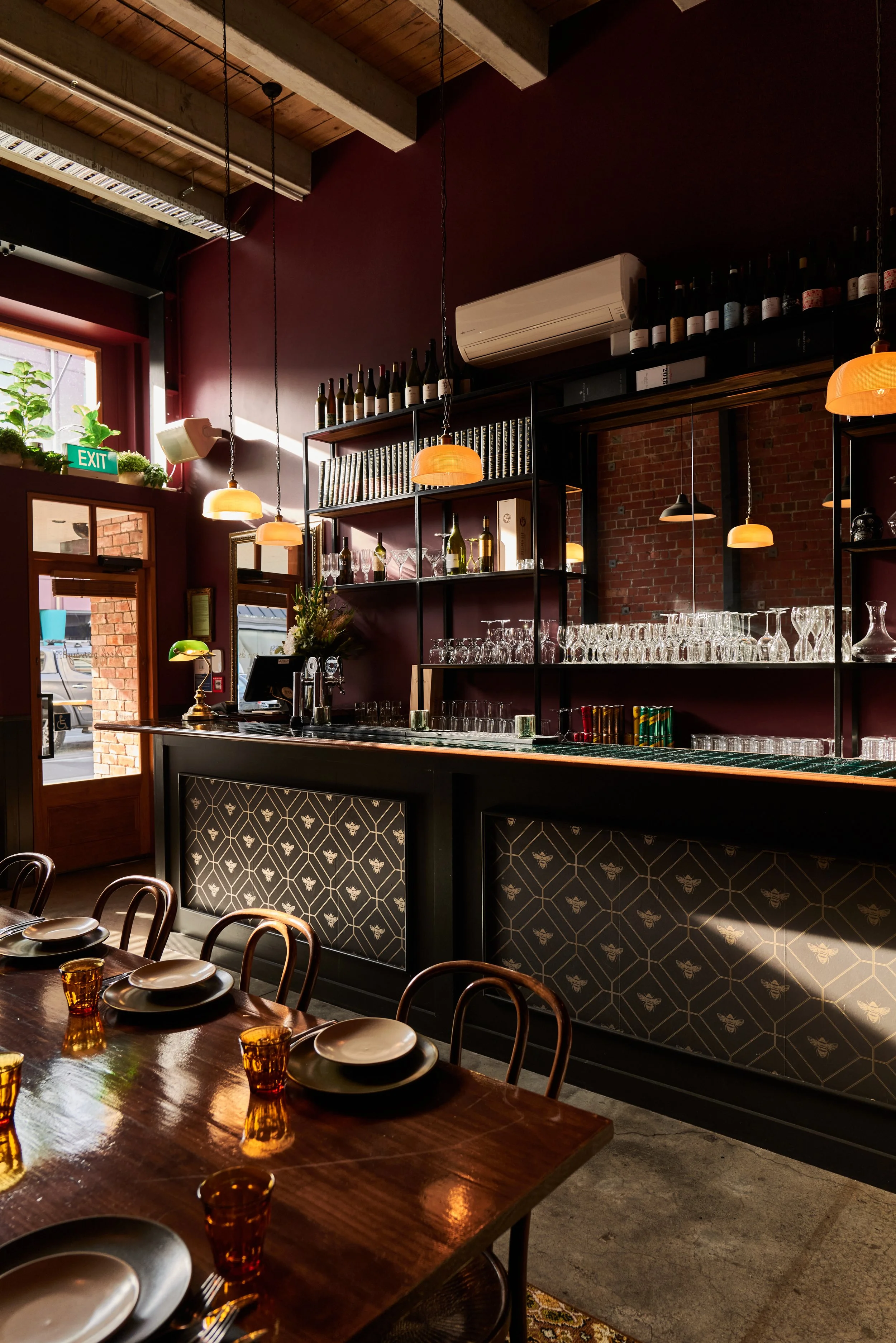 Interior of a restaurant or bar with a wooden table set with plates and glasses, dark patterned paneling, a bar counter with glassware, shelves with bottles and books, burgundy walls, windows with plants, and hanging light fixtures.