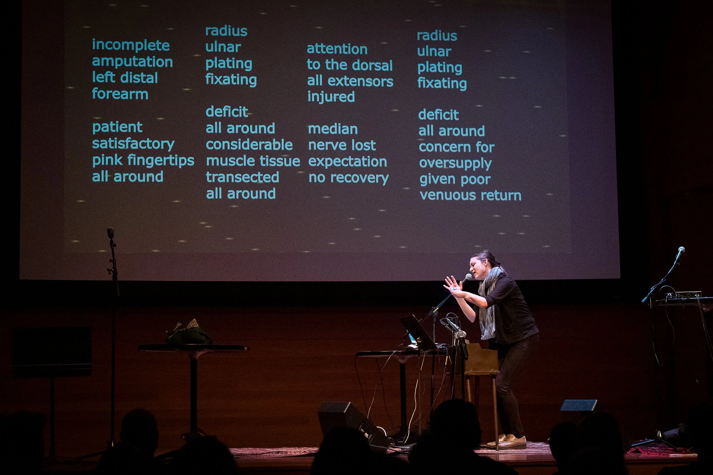 NYFOS Festival, Rubin Museum, New York, NY. image description: I sing into a mic onstage, with projection behind me displaying the lyrics.