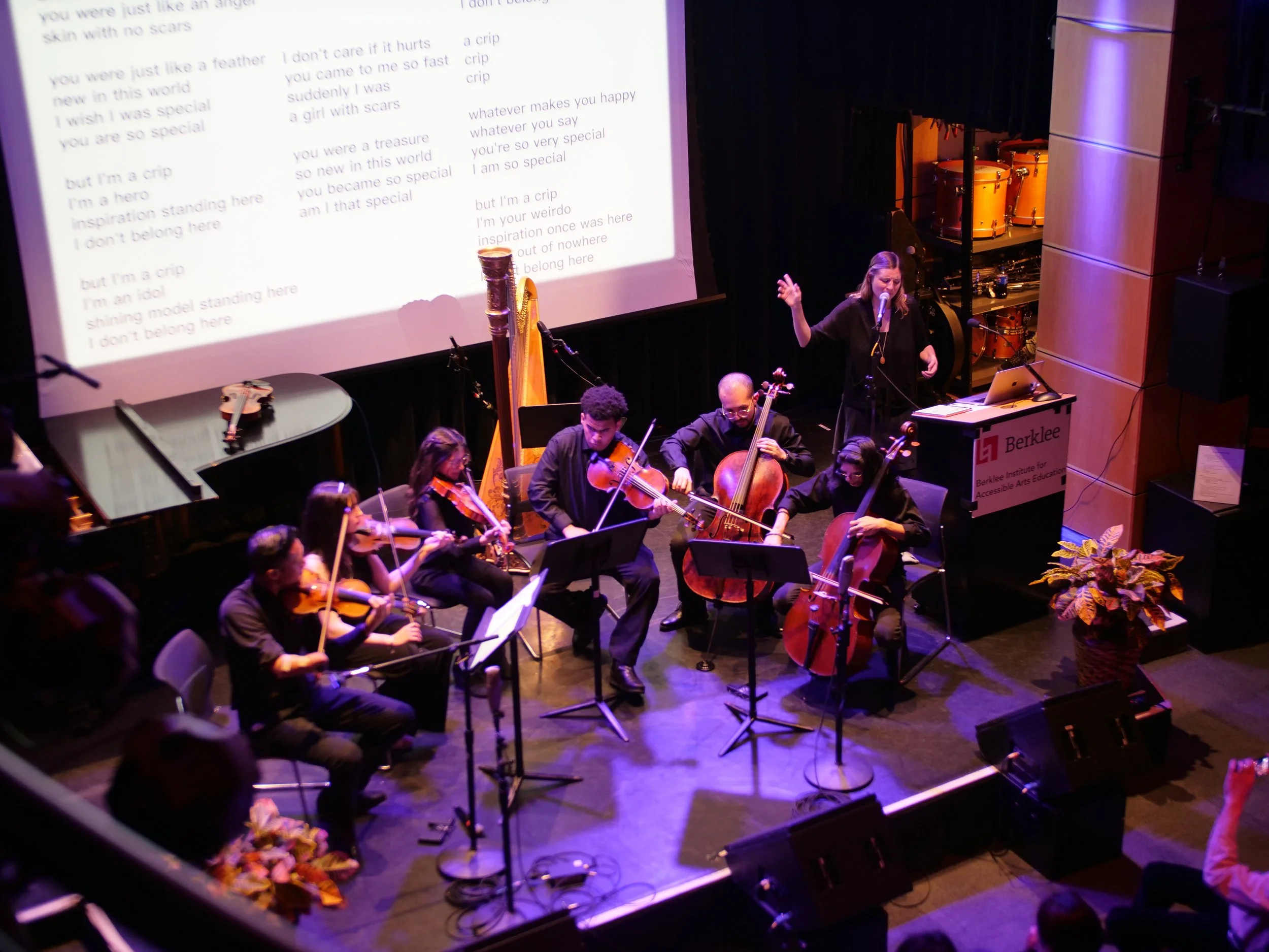Music Inclusion Ensemble, Berklee College of Music, Boston, MA. image description: I perform at a lectern with projection behind me displaying lyrics, alongside the Music Inclusion Ensemble (mostly string instruments/players).