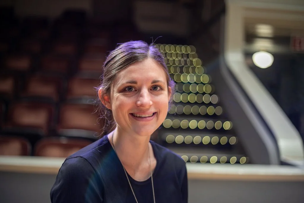 image description: I smile into the camera, with seating of a concert hall behind me. I am a white female with brown hair, and my hair is tied up in a bun.