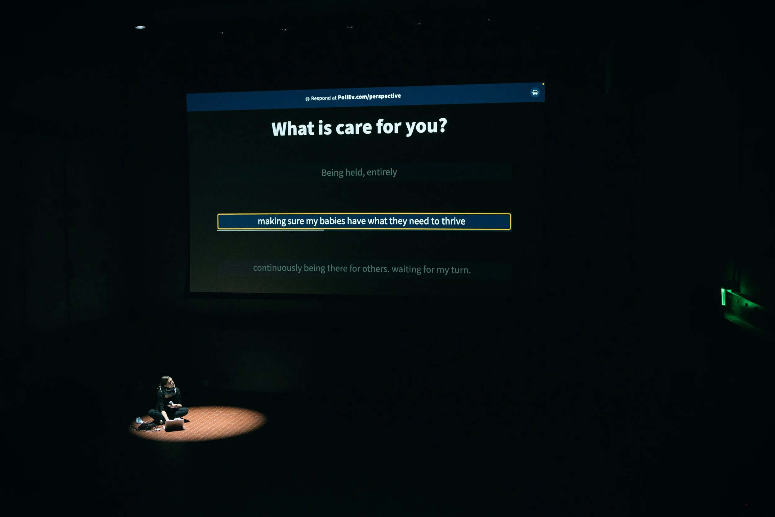 INVERSE Performance Art Festival, The Momentary, Bentonville, AR. image description: I perform in a dark space, and my performance area is spotlit. I am looking onto a projection screen with the question "What is care for you?" and the answer: "makin