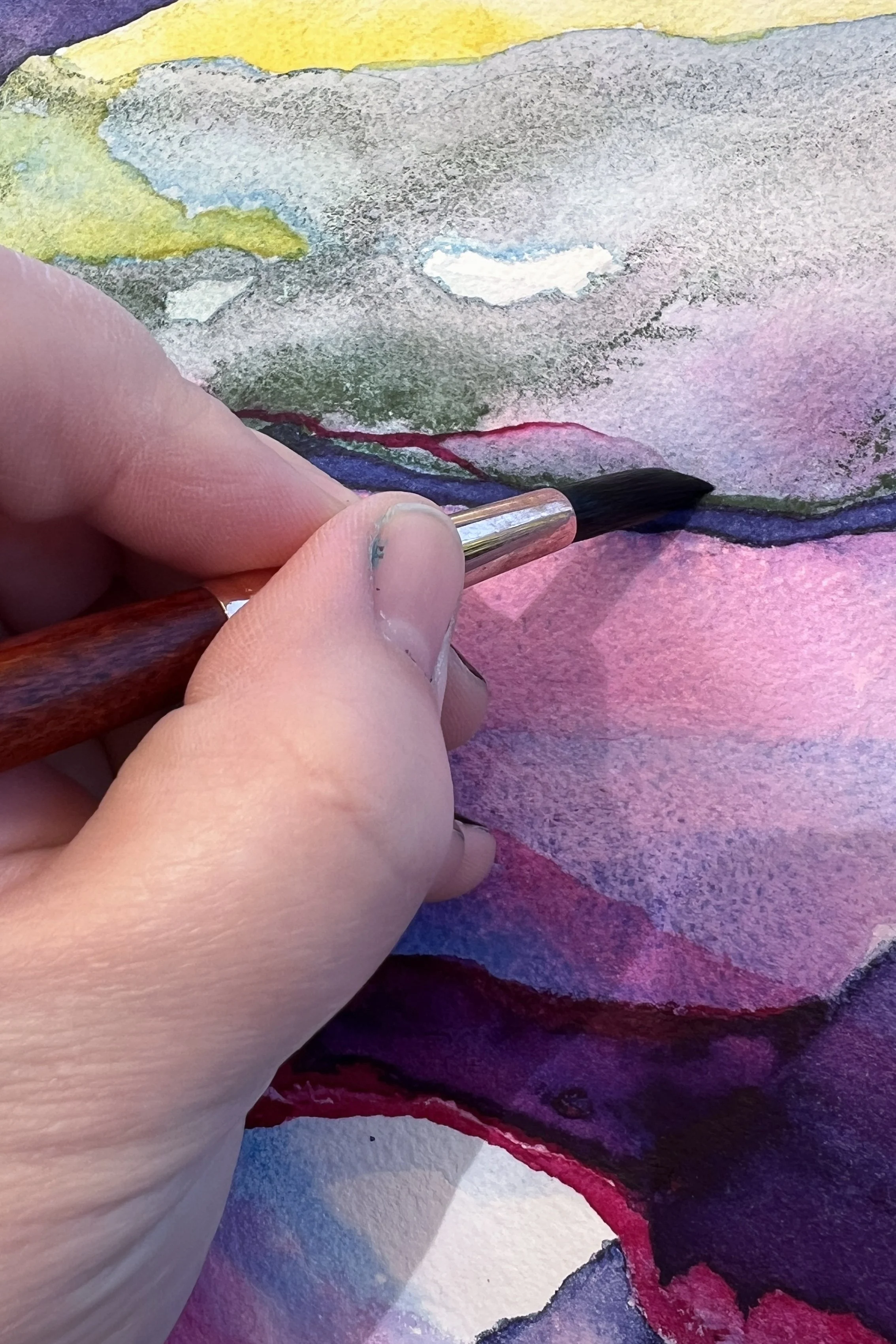 Close up of artist Mindy Wara's hand painting an original watercolor in purples, blues, pinks, greens, and yellows