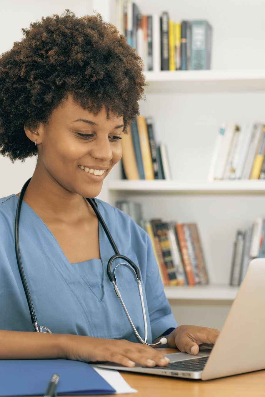 A female healthcare worker wearing scrubs and a stethoscope, smiling and working on a laptop in a medical office or library setting.