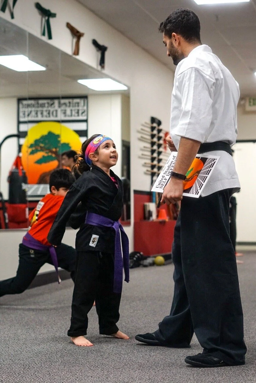 A karate instructor and a smiling kids class karate student interact in the interiors of the karate school as other karate class students practice in the background