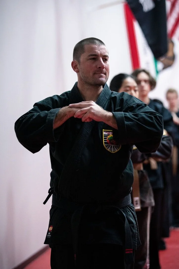 An adult karate class students at United Studios of Self defense Belmont is standing ready for class in the front position as other teens and adults do the same behind him.