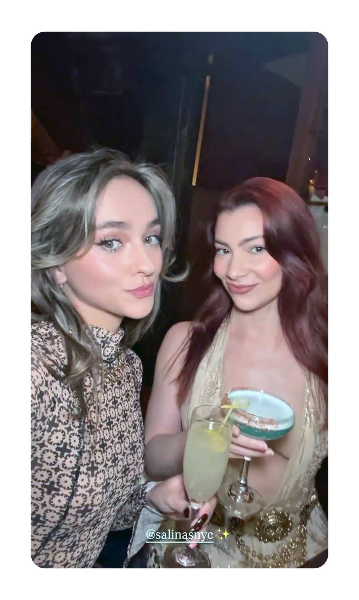Two women at a party or bar holding cocktails, one with a flute of champagne and the other with a blue cocktail in a glass, both smiling at the camera.