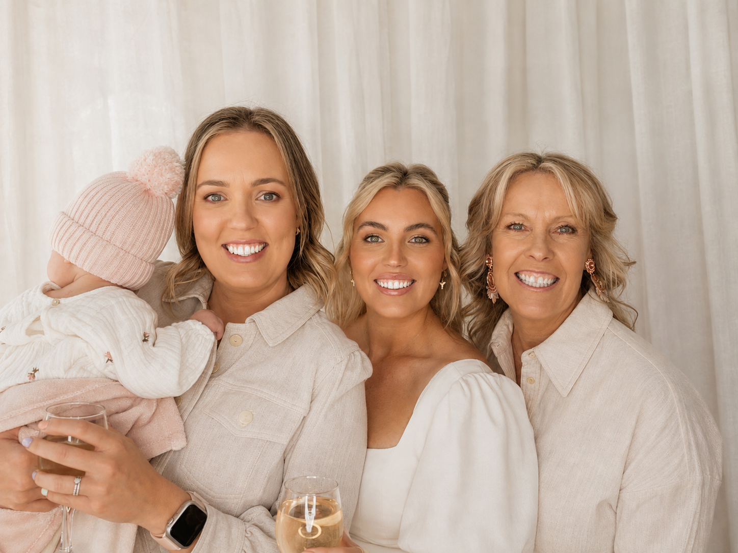 Three smiling women and a baby girl wearing a pink hat, holding glasses of champagne, standing against a light-colored curtain background.