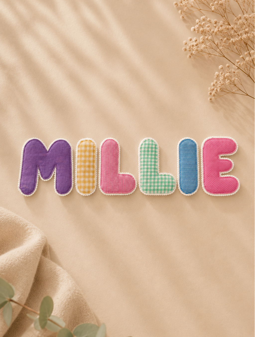 Colorful fabric letters spelling 'MILLIE' on a beige background with dried flowers in the top right corner.