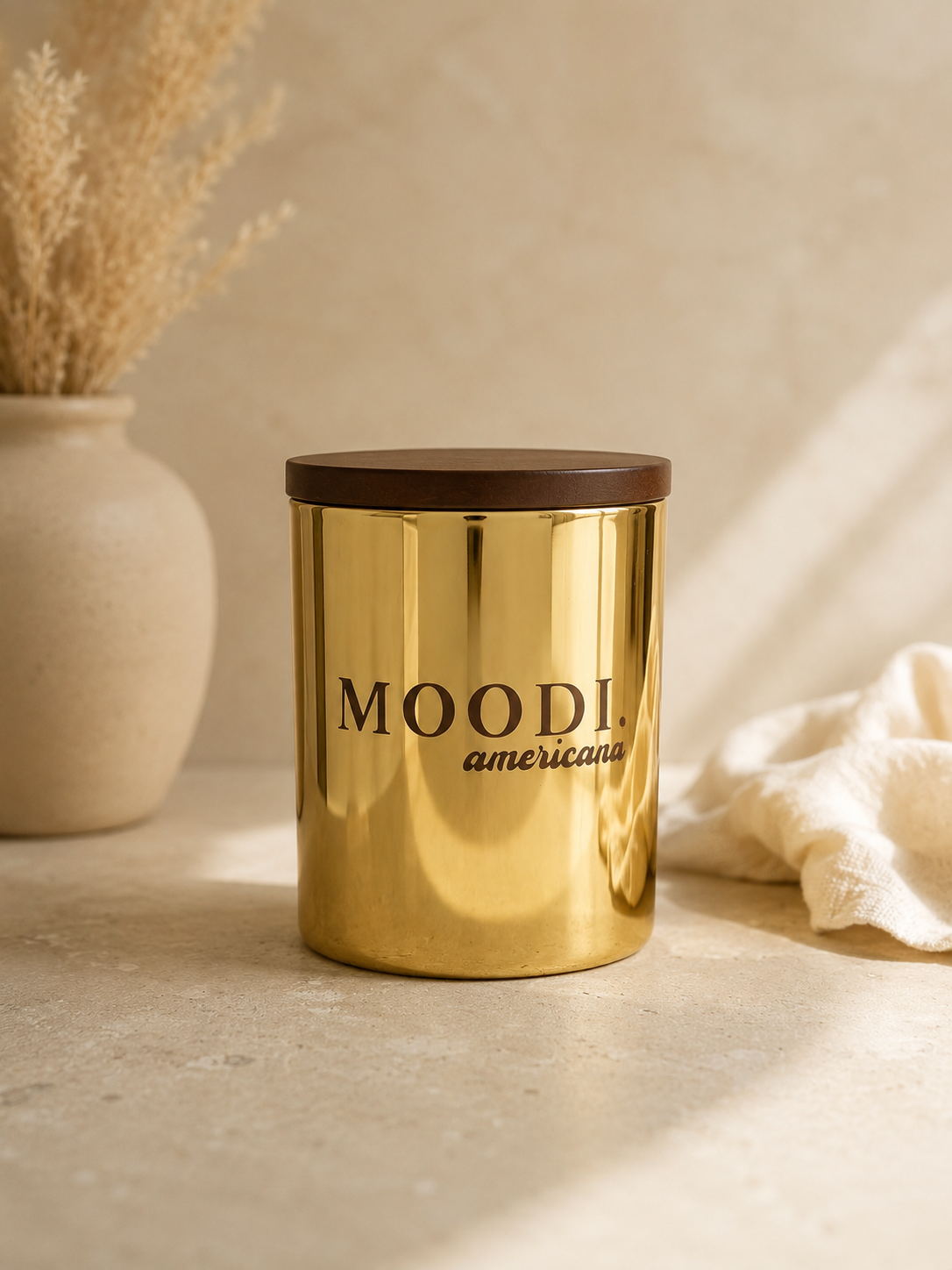 A gold-colored container with a brown lid featuring the words 'MOODI americana' on it, placed on a light-colored surface next to a beige vase with dried plants and a white cloth.