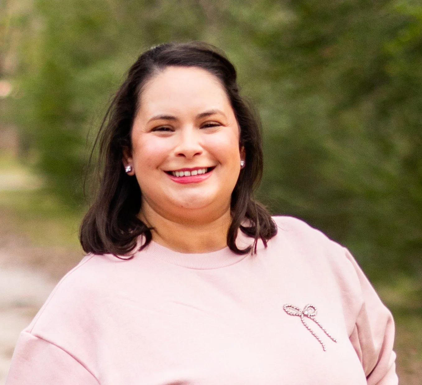 A smiling woman with dark brown hair, wearing a light pink sweatshirt and pearl earrings, stands outdoors with a blurred green background.