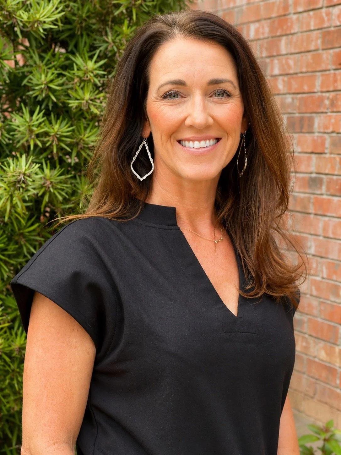 A smiling woman with long brown hair, wearing a black top, earrings, and a gold necklace, standing outdoors with green plants and a brick wall in the background.