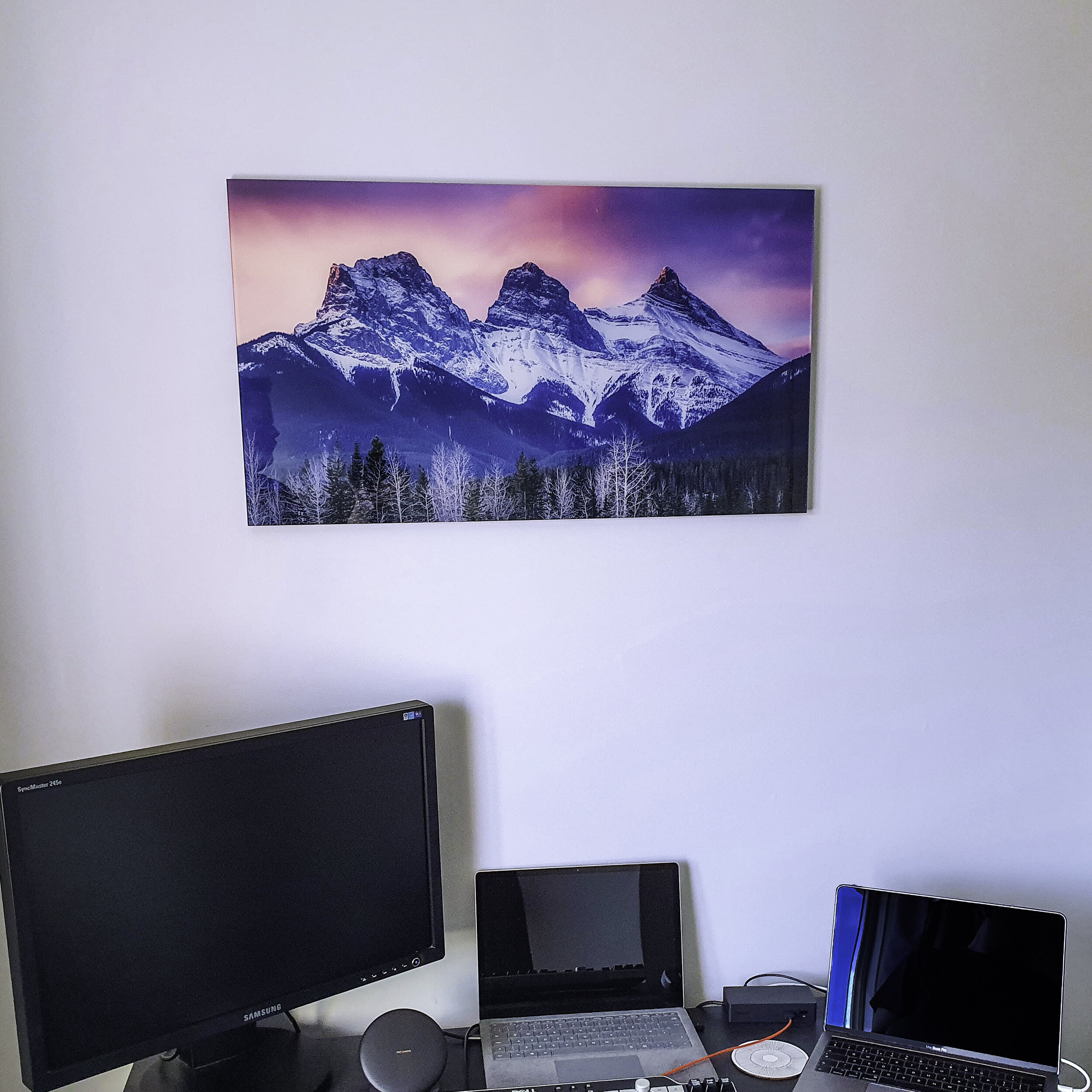 Photo of a home office with a wall-mounted landscape painting of snow-capped mountains at sunset, a large monitor, a laptop, a tablet, and a smaller laptop on a desk.