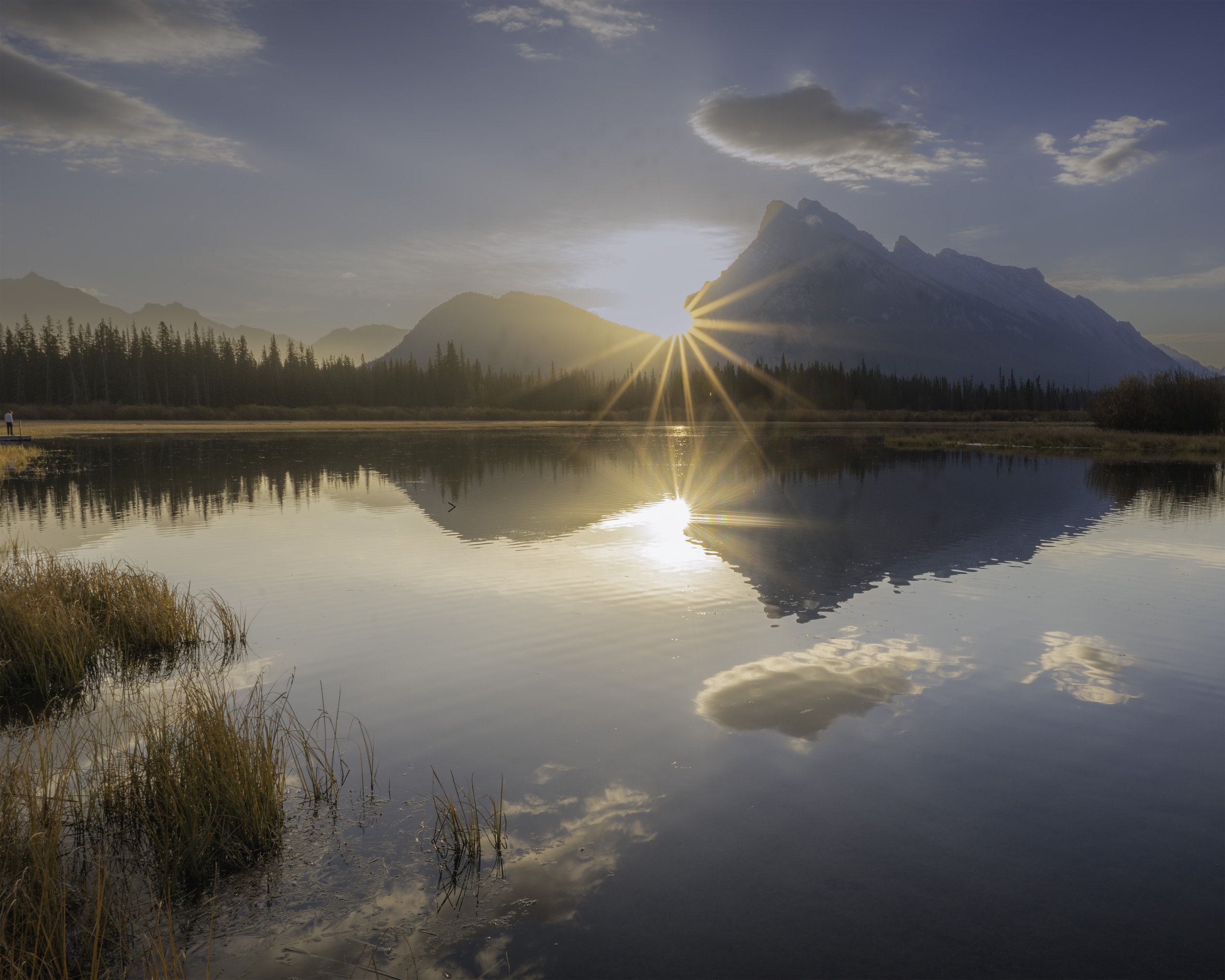 Sun setting behind a mountain range reflected in a calm lake, with trees along the shoreline and grass in the foreground.